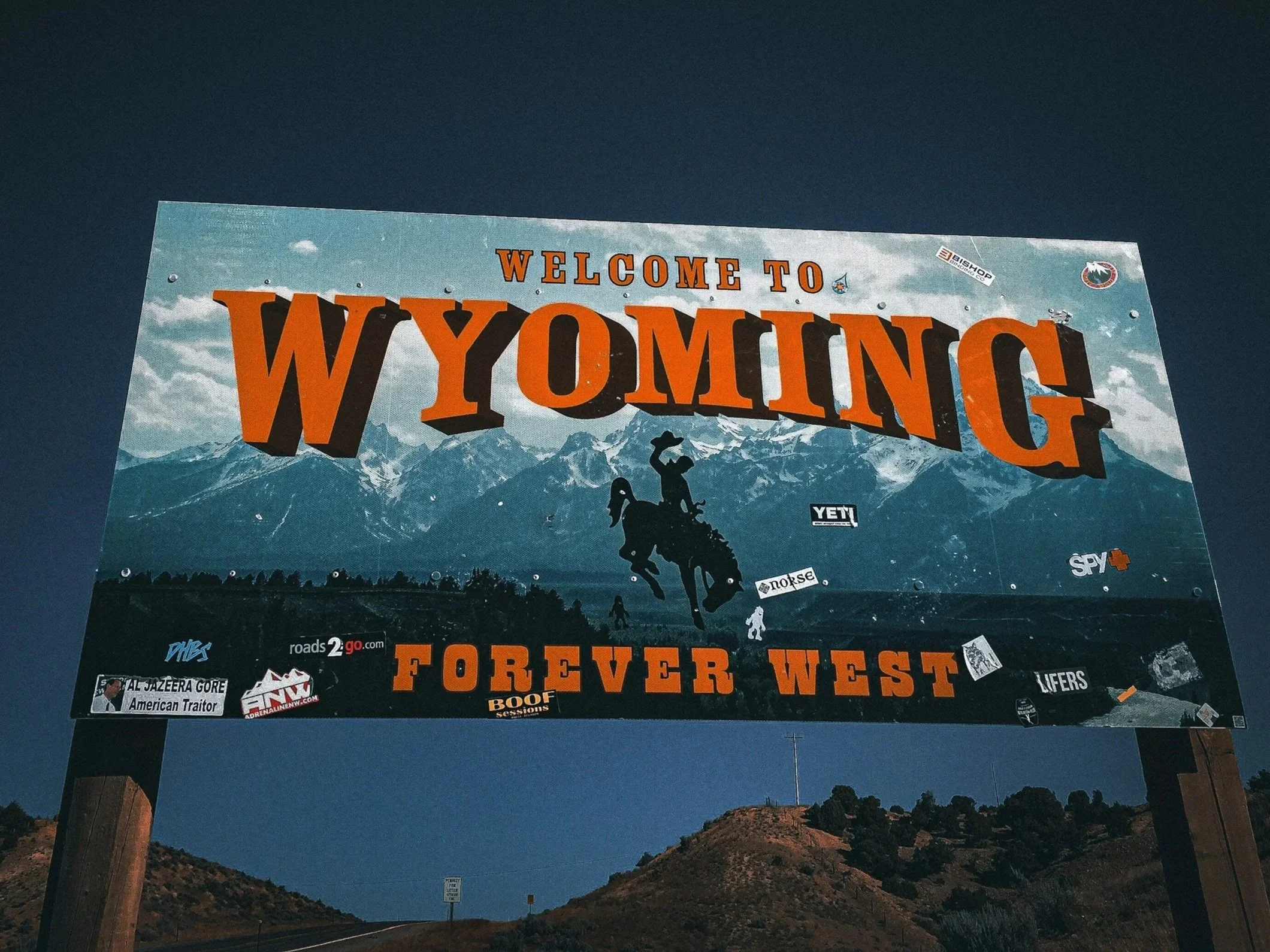 photo of a roadside billboard that says 'welcome to wyoming. Forever west' in orange font. the background of the sky is blue and you can see the night sky behind the billboard