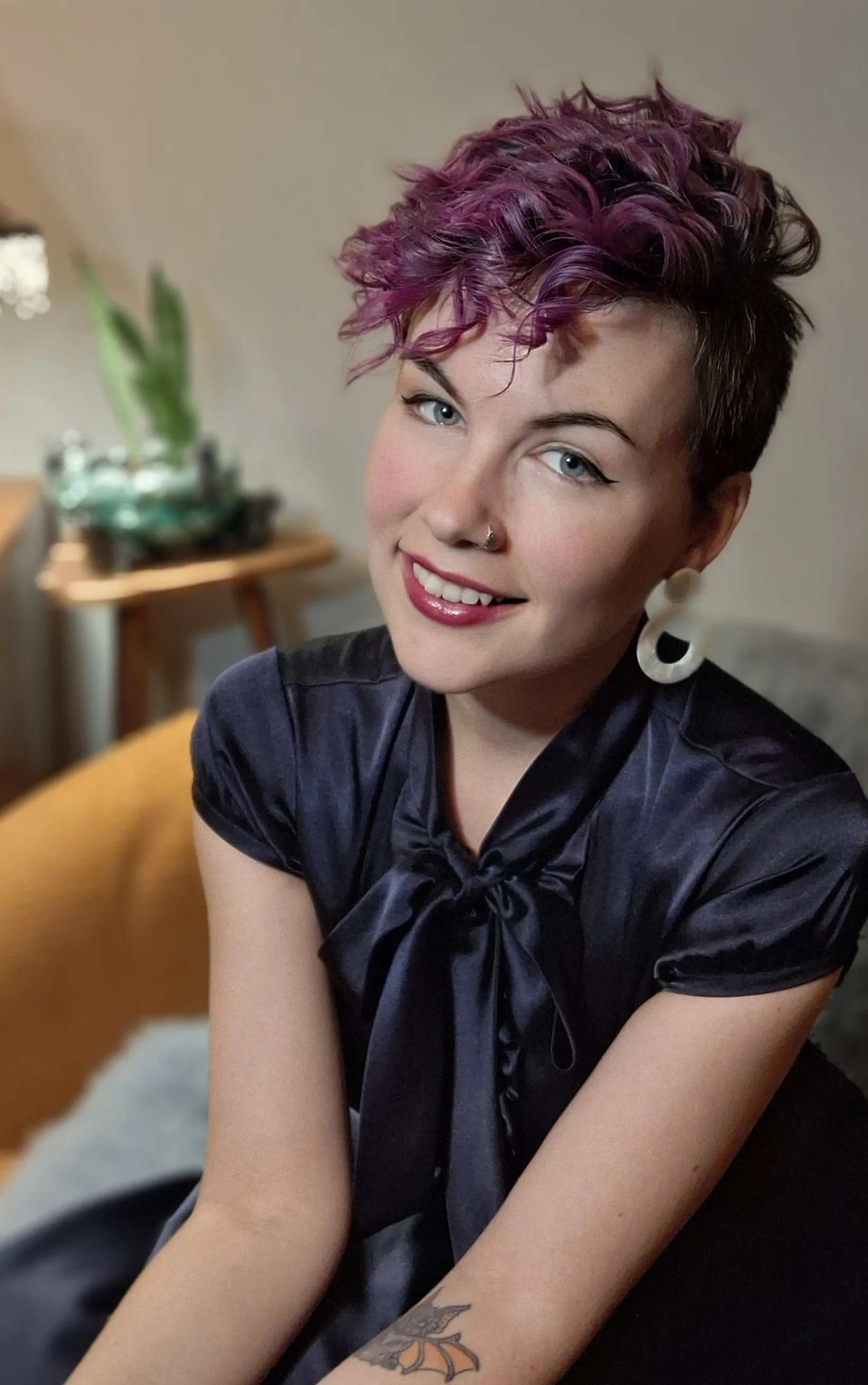 A woman with short, curly purple hair, wearing a black satin blouse with a bow tie, large white earrings, and a nose ring, sitting on a couch in a cozy living room.
