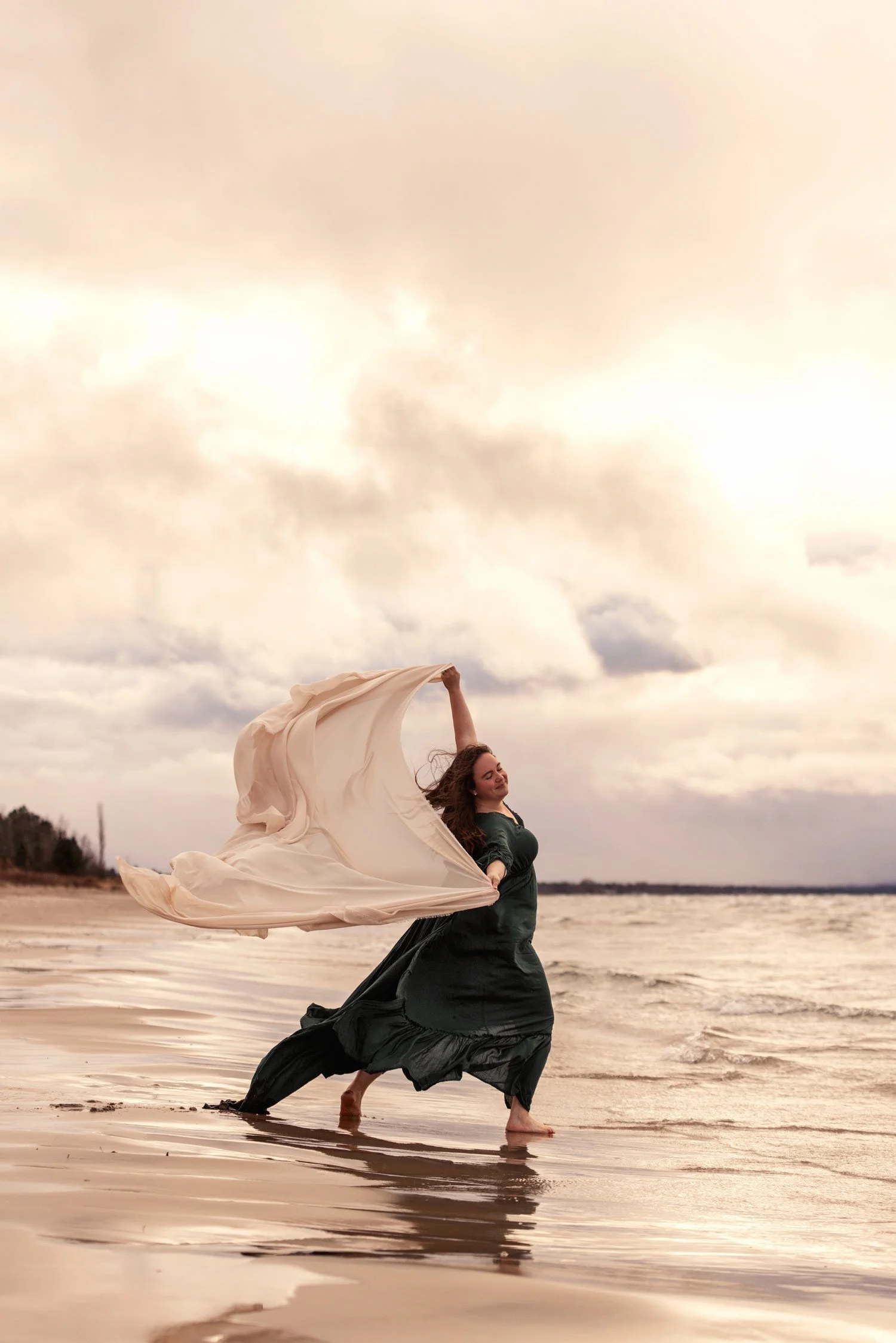 Woman in a green dress on a beach holding a flowing fabric in the wind.