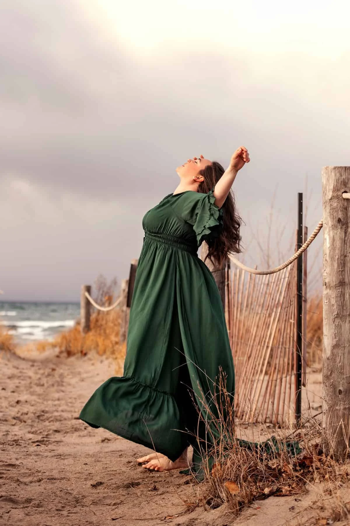 Woman in green dress standing on a beach path, arms stretched out, with ocean waves in the background.