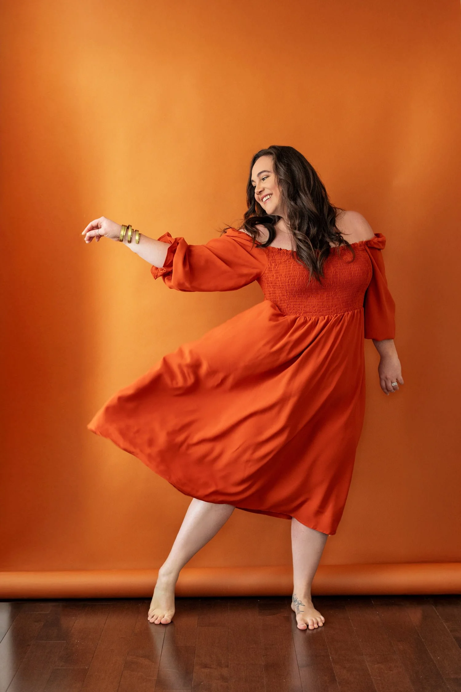 Woman in an orange dress dancing barefoot against an orange backdrop.