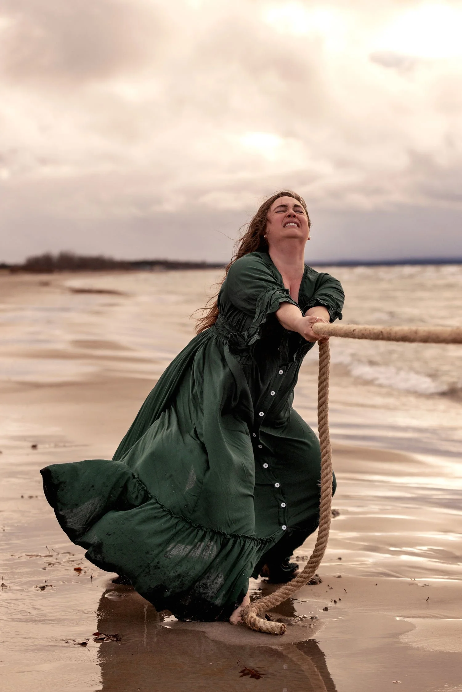 A woman in a green dress pulling a rope on a beach during cloudy weather.