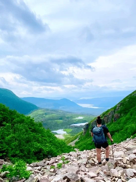 Hiker with backpack on rocky mountain trail overlooking lush valley and distant hills under a cloudy sky.