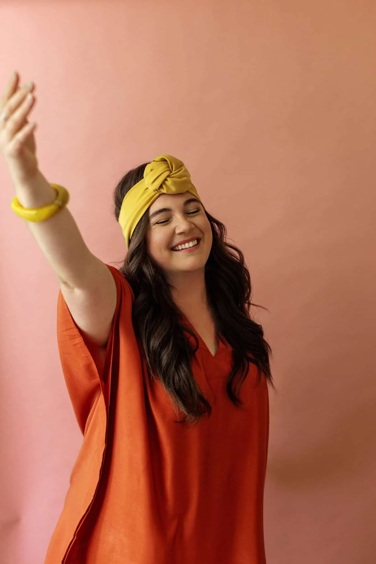 Woman smiling with closed eyes, wearing yellow headband and orange shirt against a pink background.