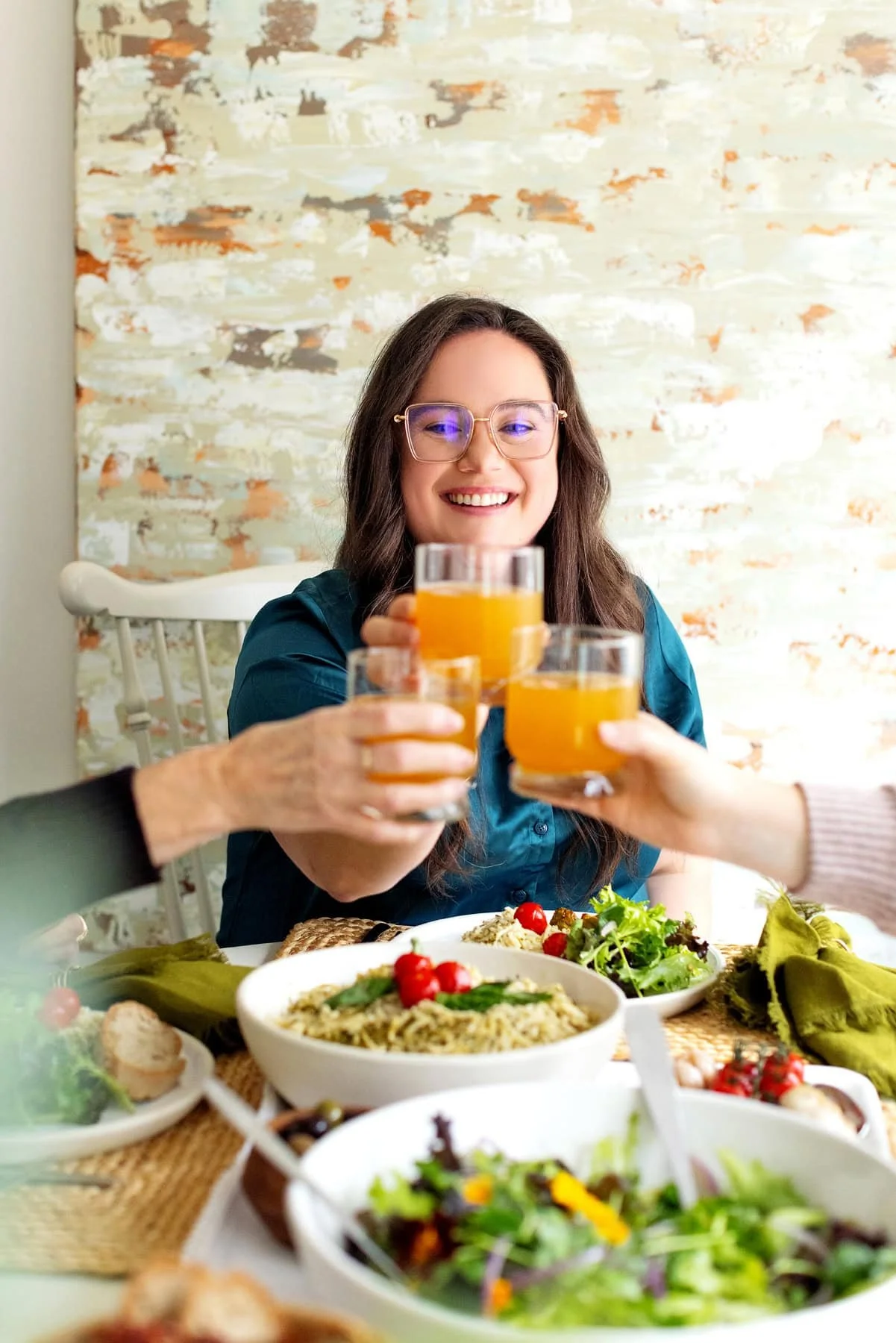 People toasting with orange drinks at a dining table with salads and pasta dishes.