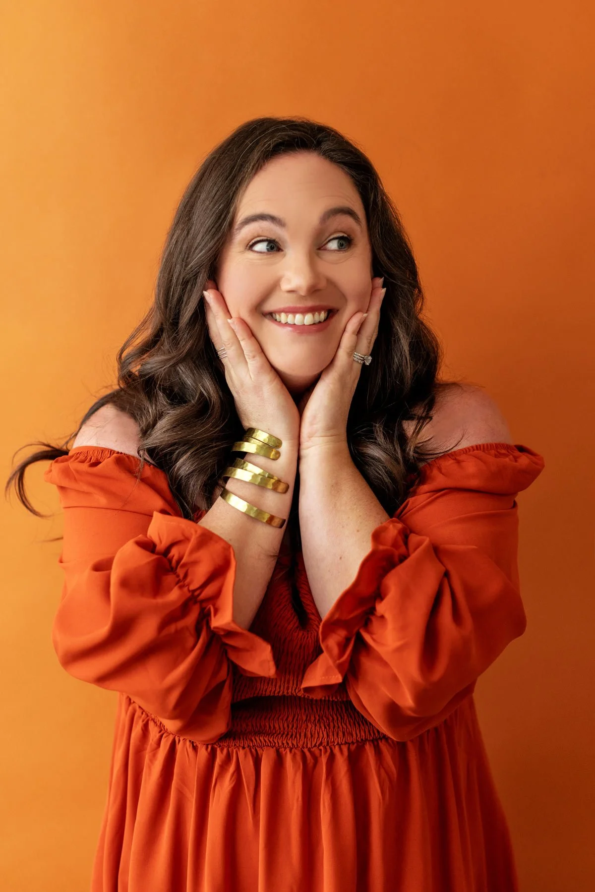 Woman in an orange dress smiling against an orange background, with hands on her cheeks and gold bracelets on her arm.