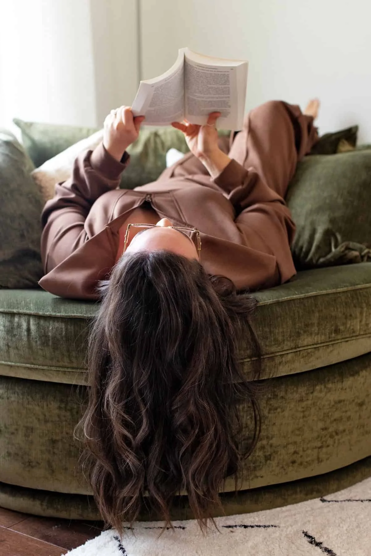 Person lying on a green couch reading a book, wearing a brown outfit with long hair hanging down.