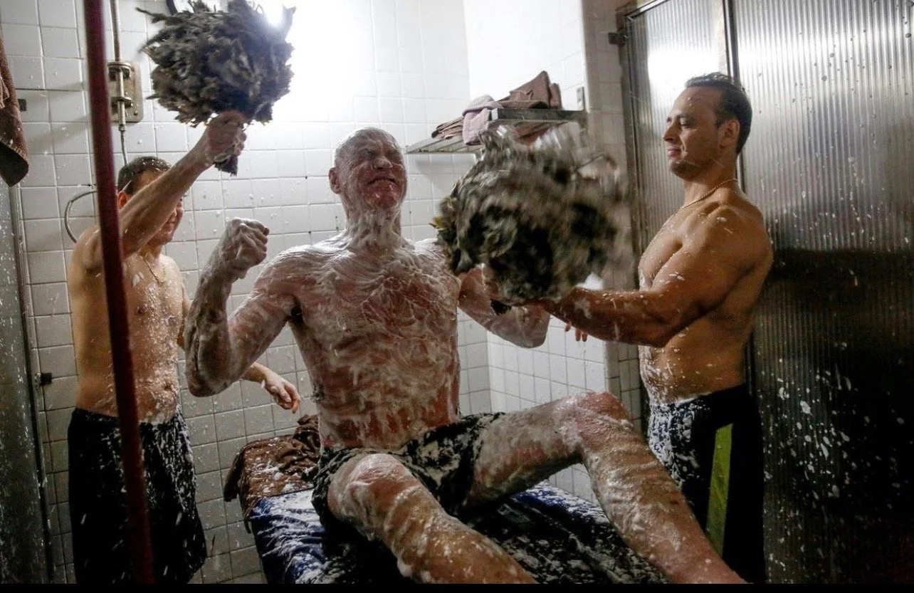 People taking a shower with soap and foam while a man sits on a bench in a shower room.