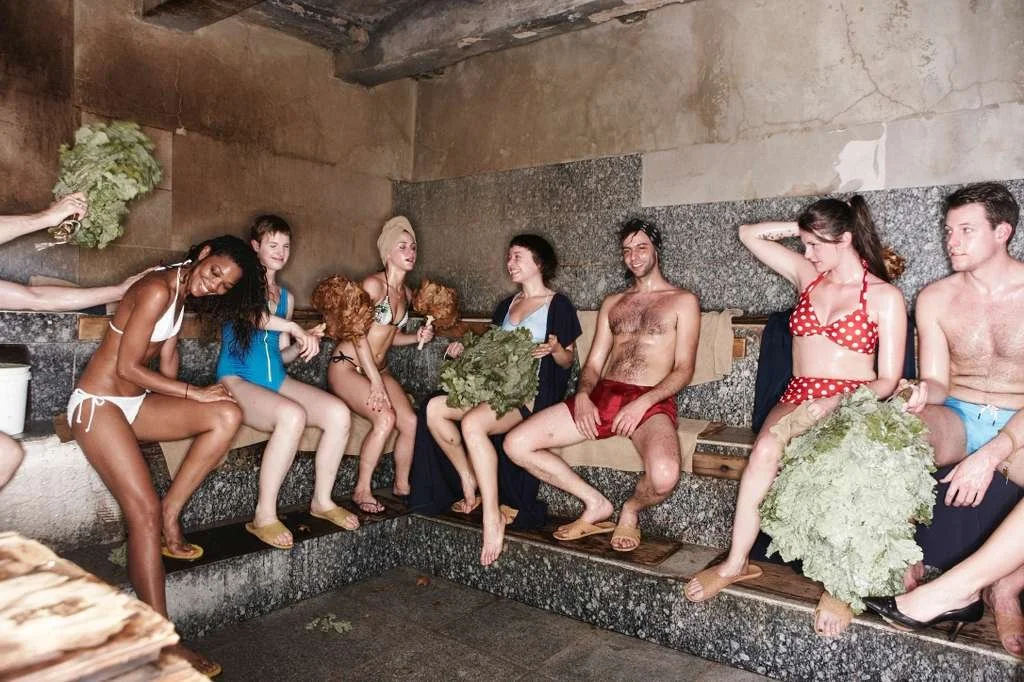 People sitting on benches in a sauna, holding leafy vegetables and engaged in conversation.