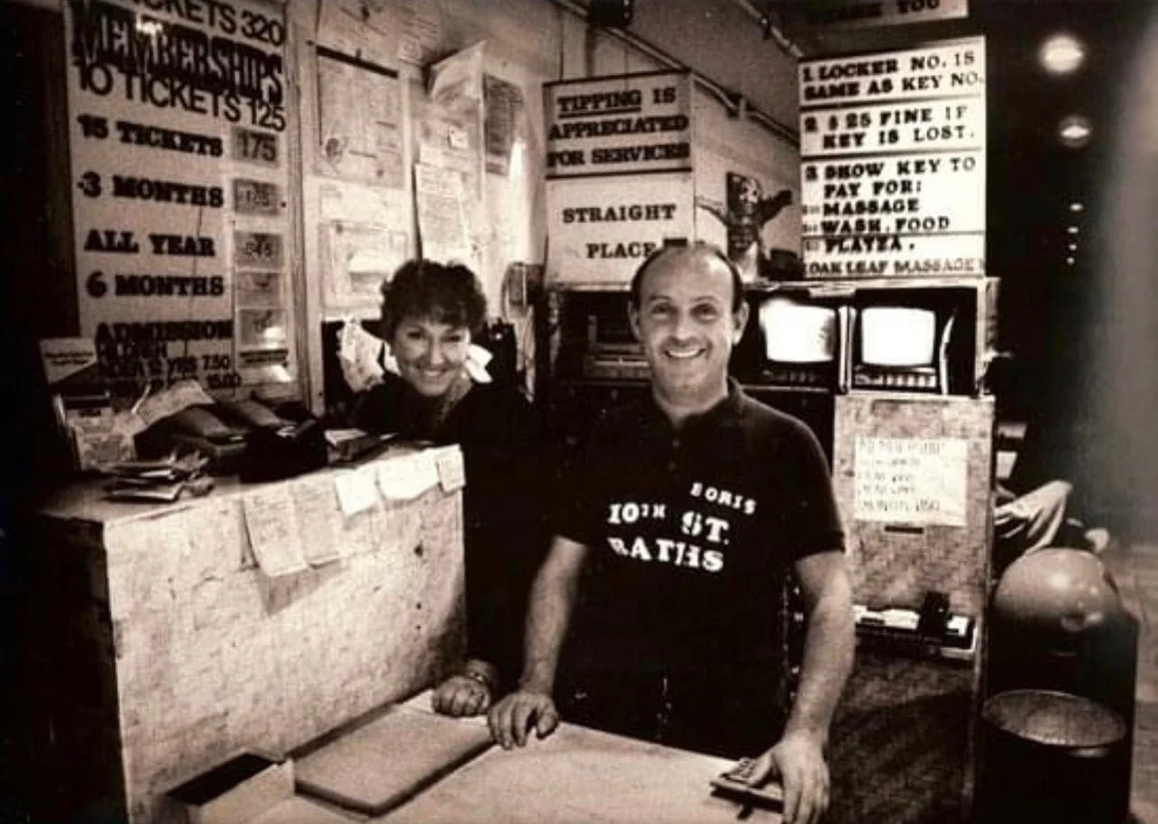 A black and white photo of a man and a woman behind a counter at a service station or store, smiling toward the camera. The man is wearing a shirt that says 'LORE 10th ST. RATES' and the woman is standing behind the counter surrounded by signs postin