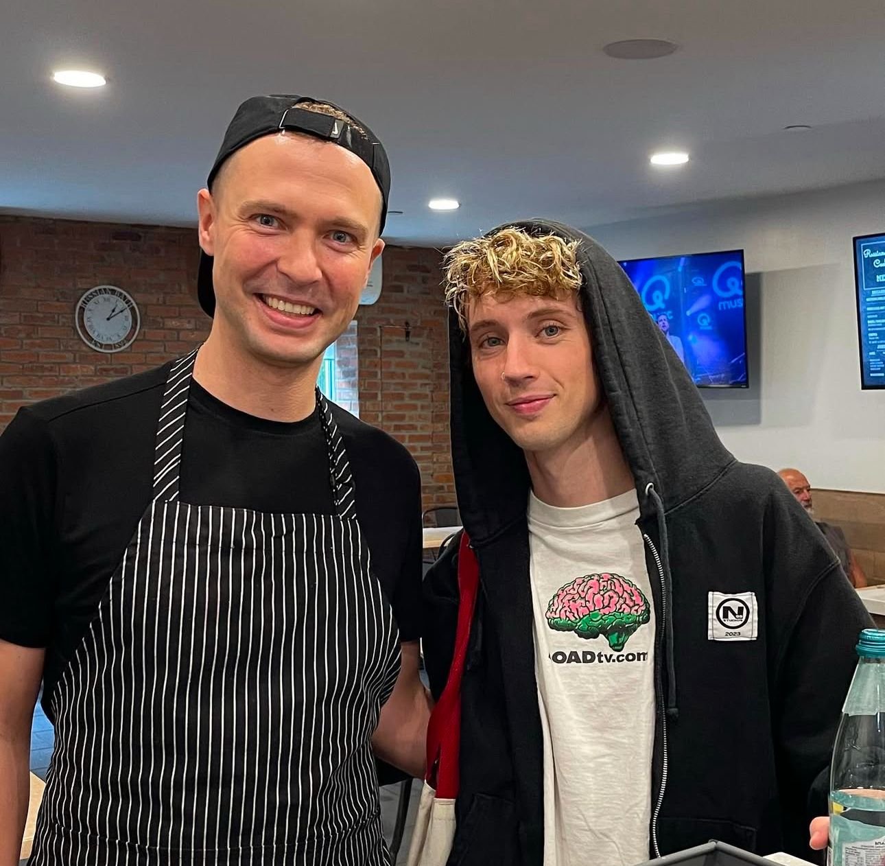 Two young men posing together in a restaurant. The man on the left is wearing a black baseball cap backwards, a black T-shirt, and a black and white striped apron. The man on the right has curly blonde hair, a nose piercing, and is dressed in a black