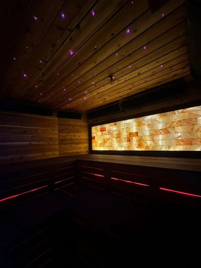 A dark room with wooden walls and ceiling, illuminated by purple star-like lights on the ceiling and a glowing amber wall. There are black benches with red lighting underneath.