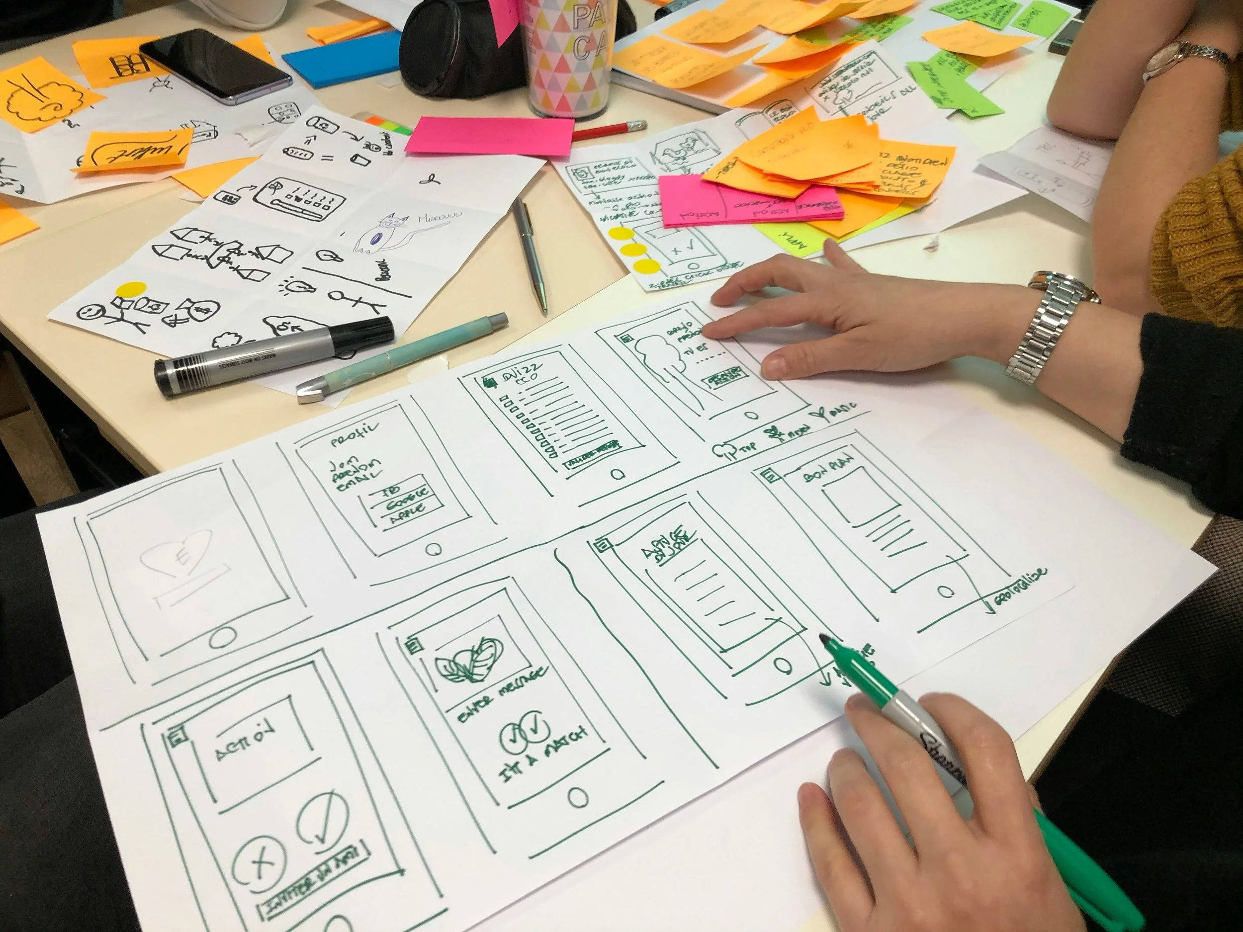 A brainstorming session at a desk with sketches of mobile app designs, colorful sticky notes, pens, and paper. Someone is working on wireframes for a mobile phone app layout.