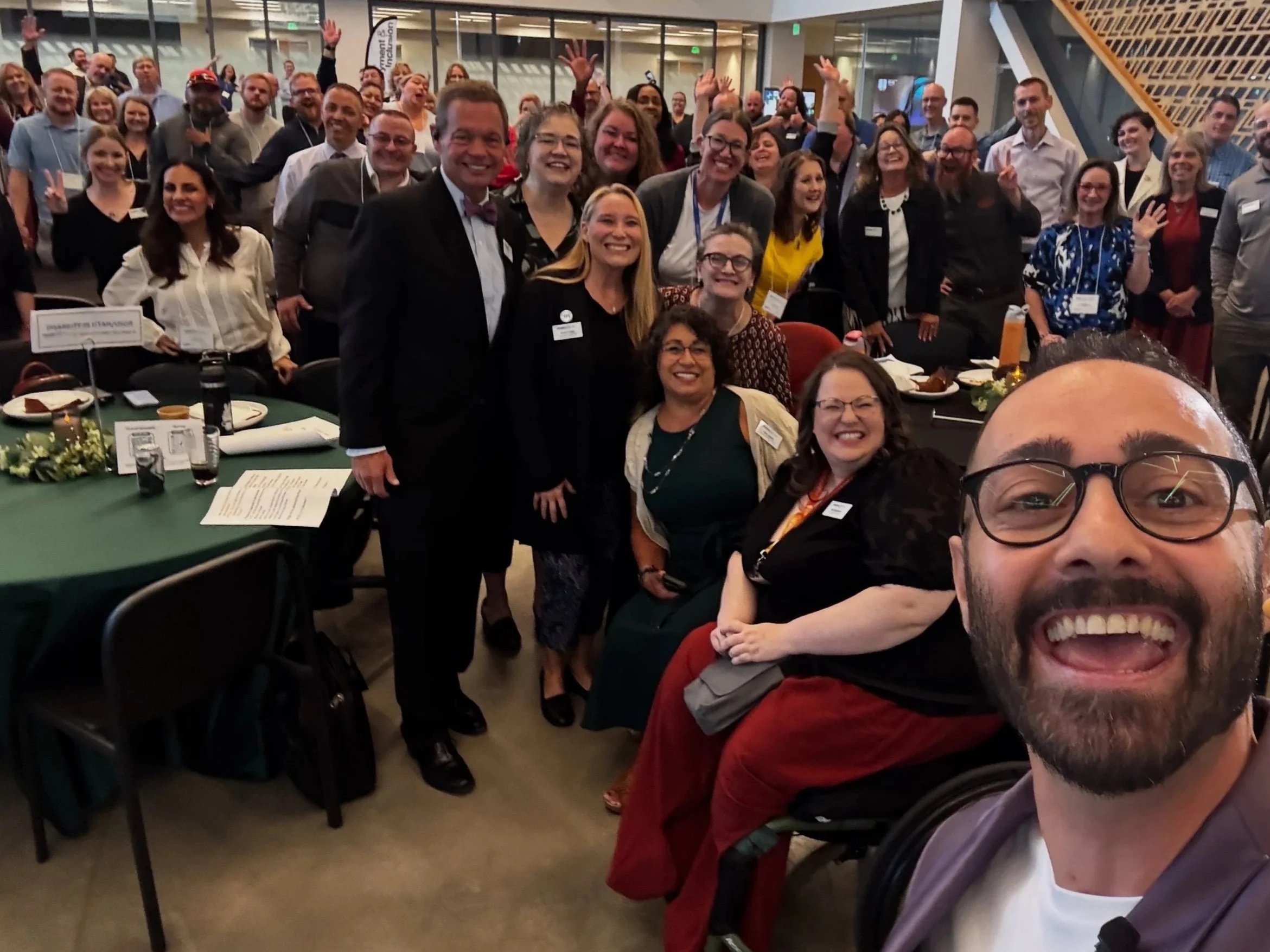 Group of people at a conference or event, smiling and posing for a selfie, with tables set for a meal in the foreground.