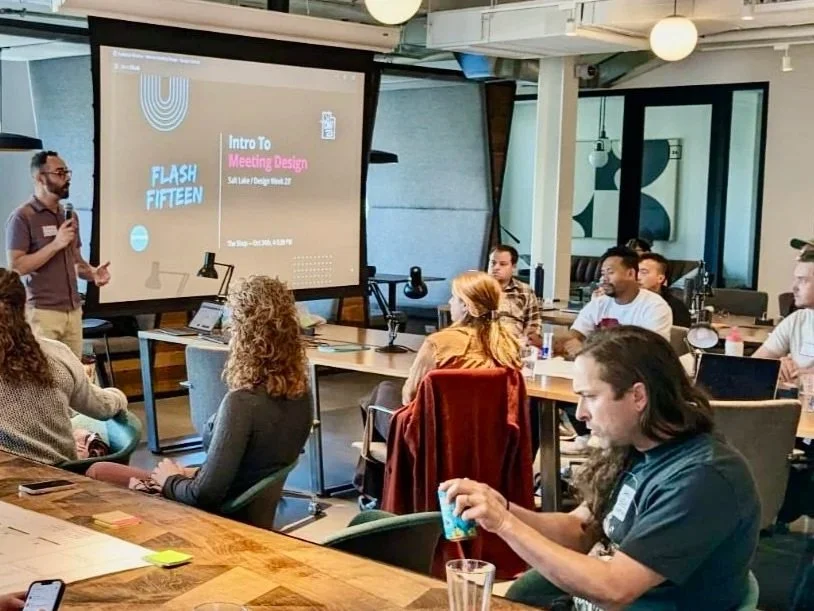 A presenter stands at the front of a room giving a presentation to a group of adults seated at tables. The presentation slide on the screen reads "Intro to Meeting Design" and "Flash Fifteen." The room has a modern, industrial decor with exposed beams and a large window.