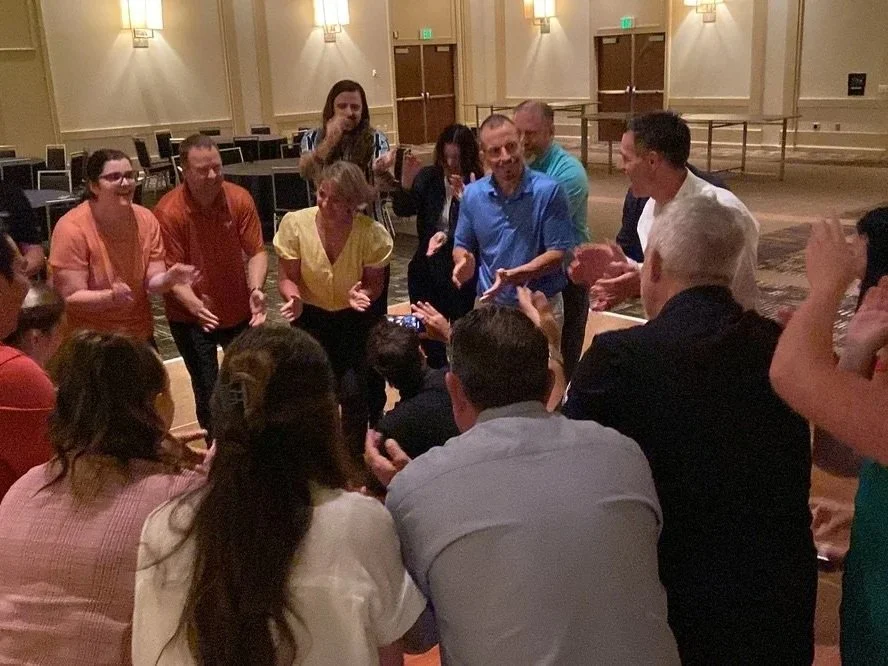 A group of people gathered together in a circle at an indoor event, some clapping and others taking photos, with a mostly empty conference room in the background.