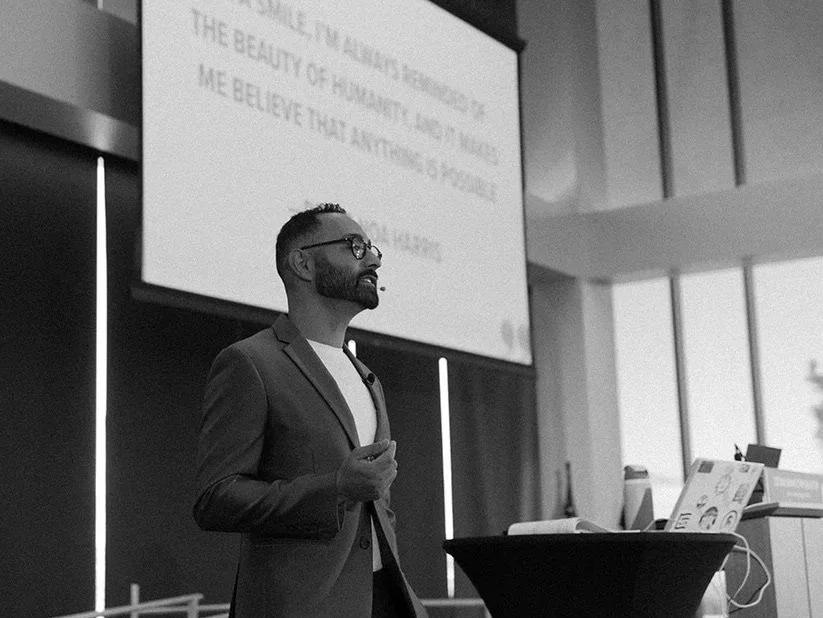 A man in glasses and a blazer giving a presentation in a conference room with a large screen displaying text behind him.