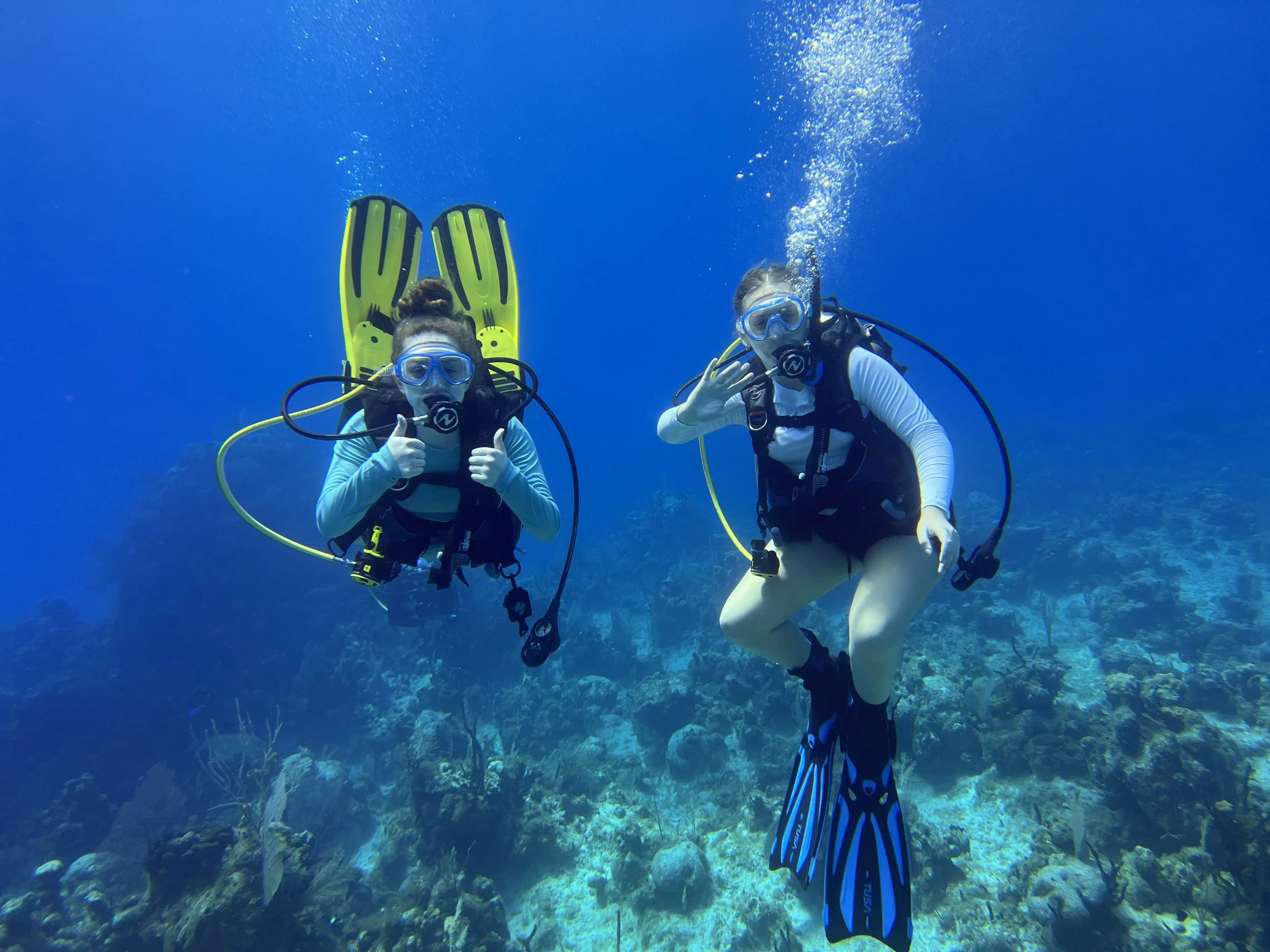 Two scuba divers underwater, giving thumbs up and waving, with clear blue water and coral reef below.
