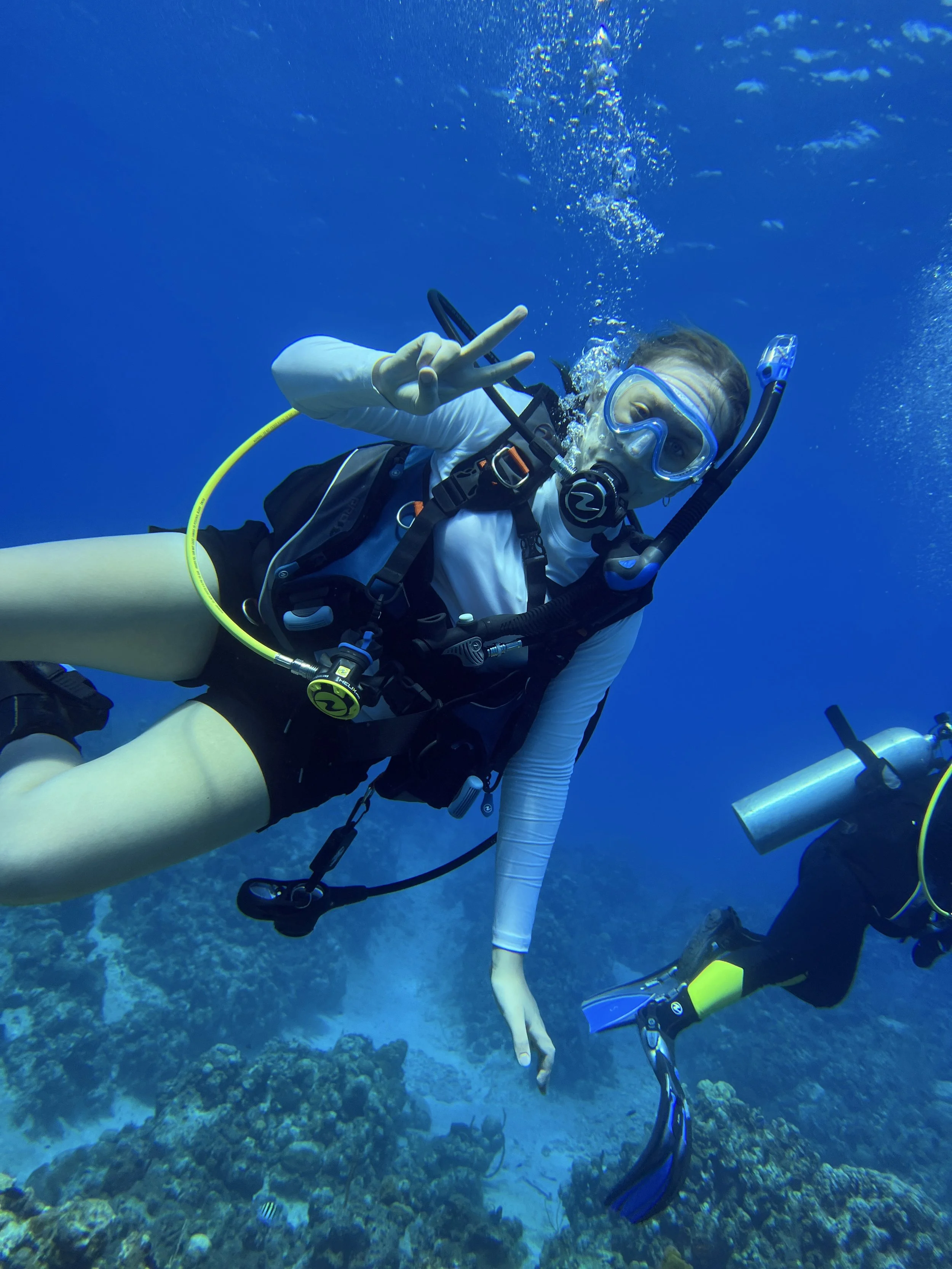 Person scuba diving underwater, wearing diving gear, making a peace sign with one hand, and floating above coral reef and fish.