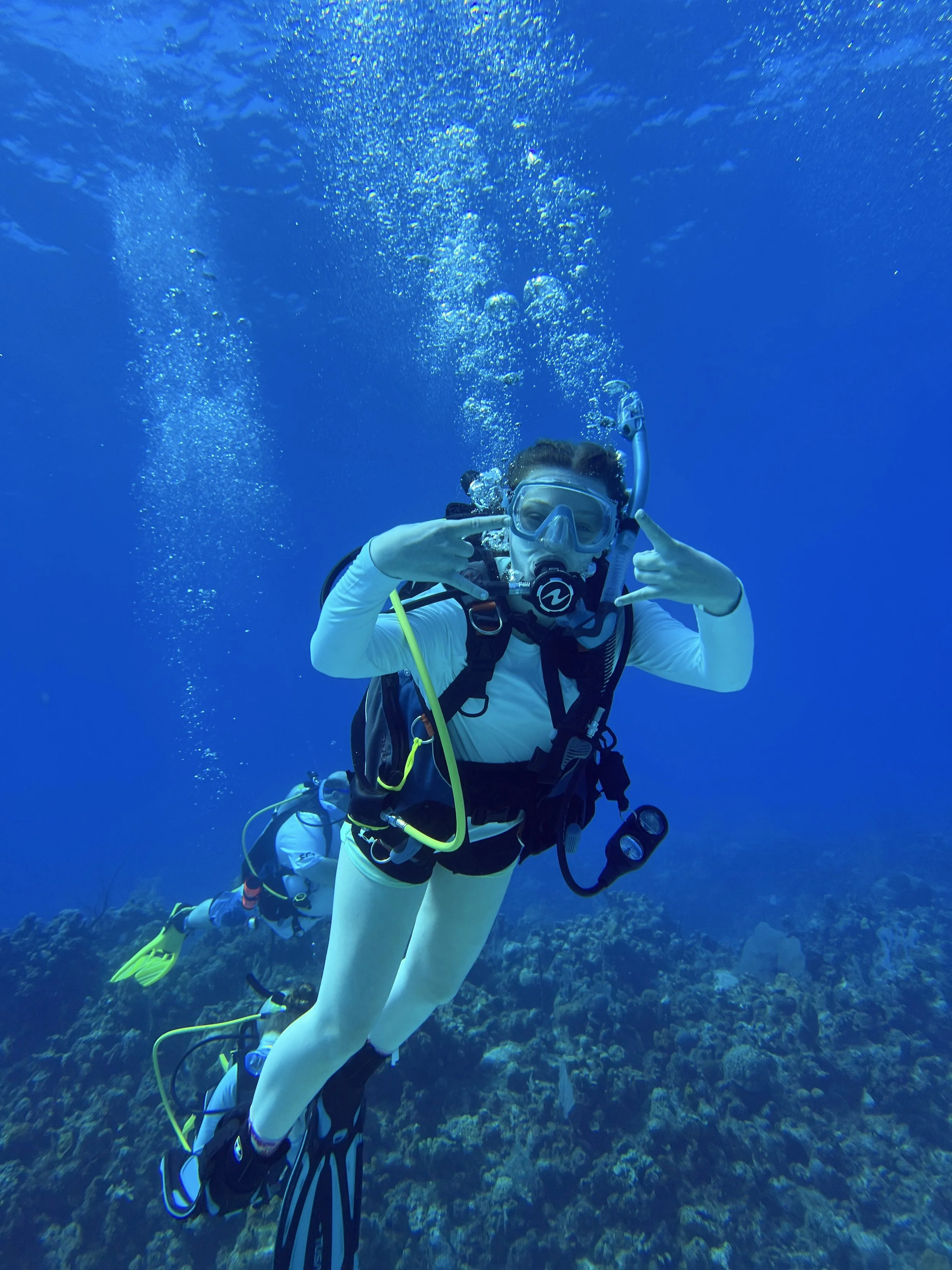 Person scuba diving underwater, wearing a mask and wetsuit, surrounded by blue water and coral reef.