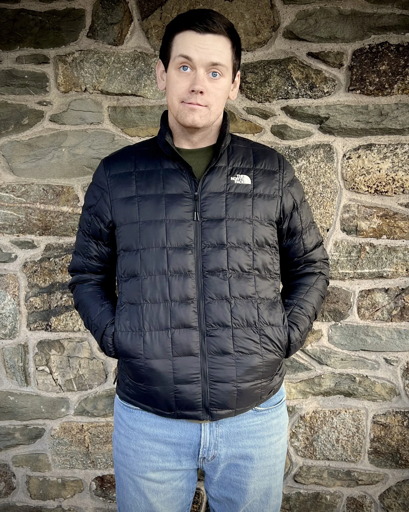 Jonathan Schimpf in a black jacket and jeans standing in front of a stone wall