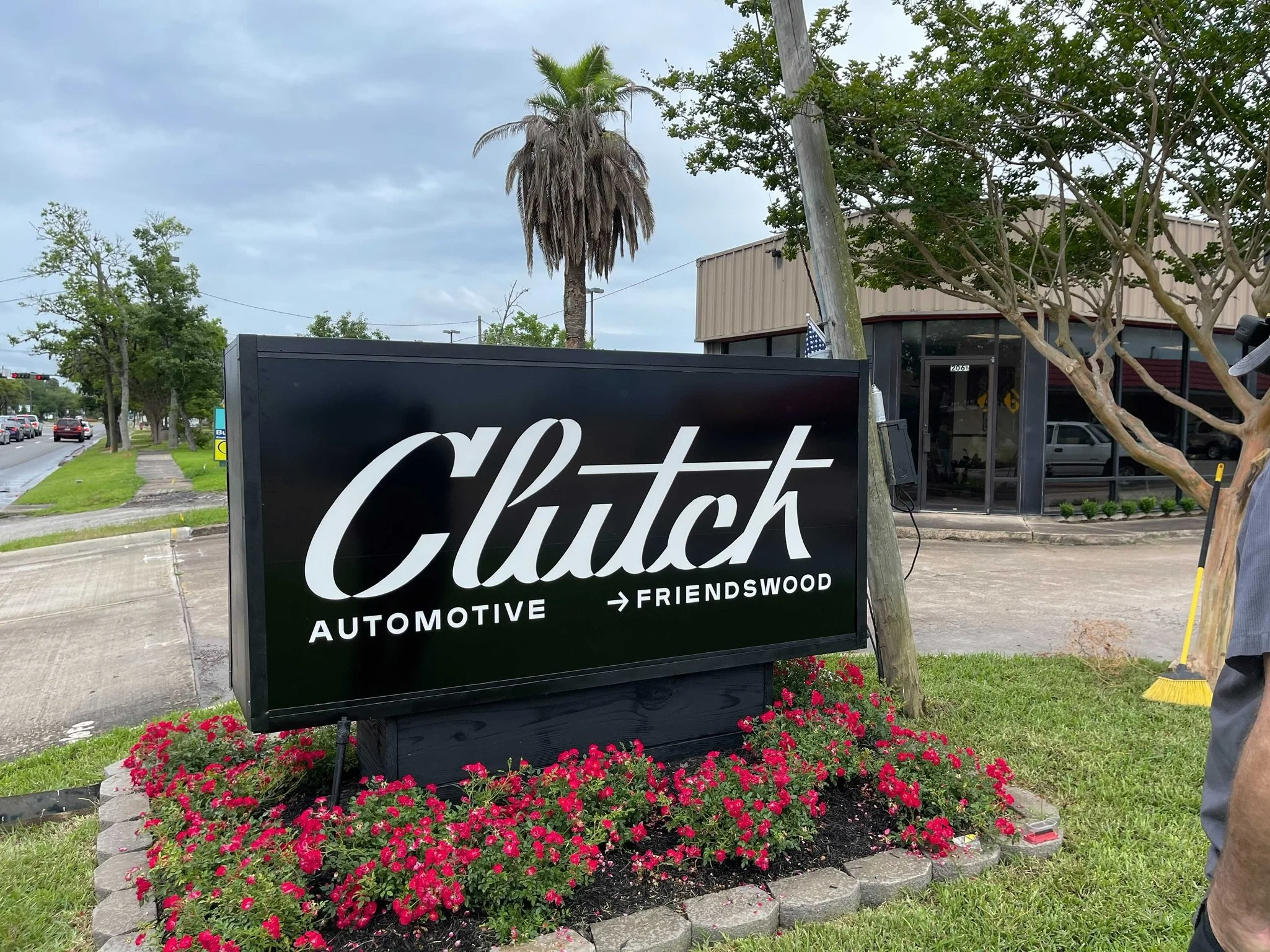 Sign for Clutch Automotive in Friendswood, with a black background and white cursive lettering, surrounded by pink flowers and a landscaped flowerbed. In the background, a street with cars, trees, and a building with beige siding and a glass window.