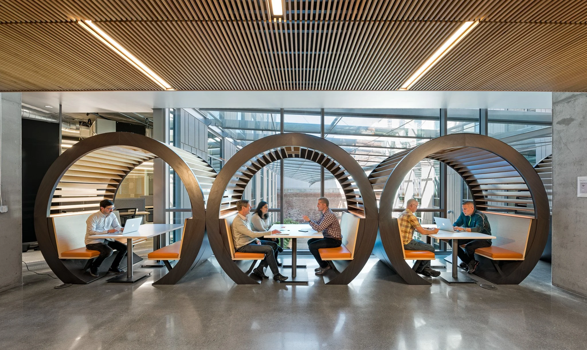 Modern office space with three round booths made of wood, each containing two people working on laptops and having discussions. Large windows let in natural light.