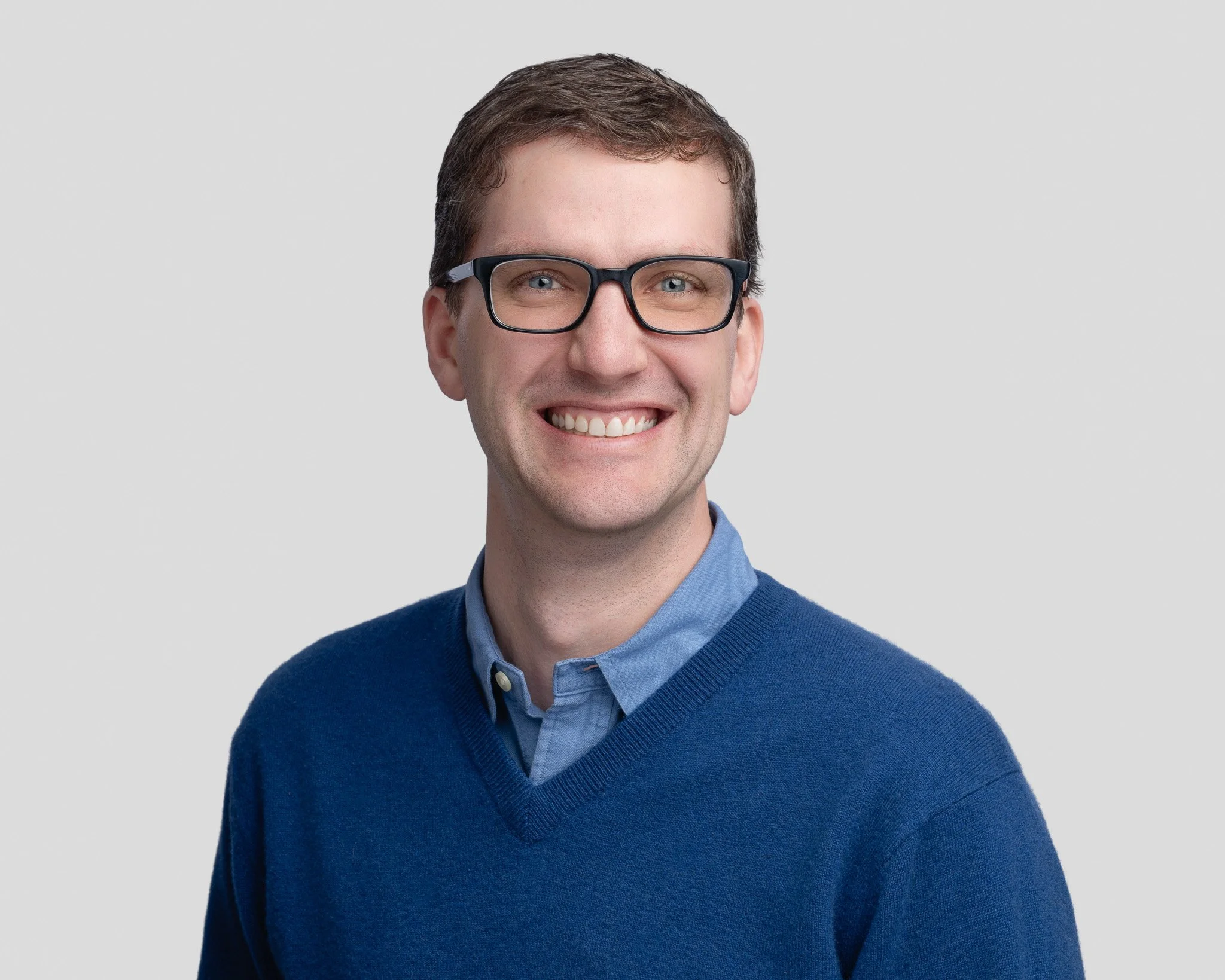 A young man with short brown hair, glasses, and light skin, wearing a blue sweater over a button-down shirt, smiling against a plain gray background.
