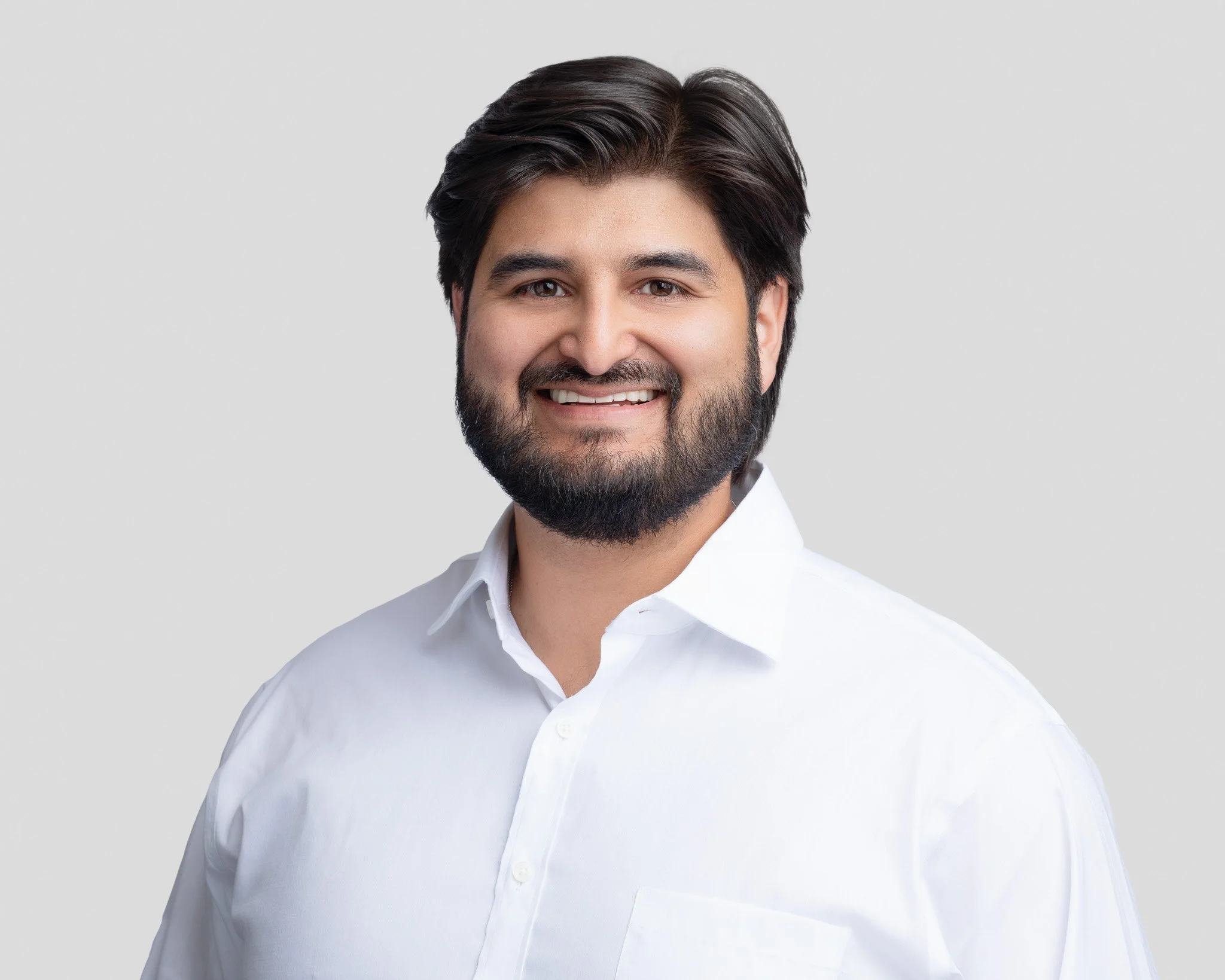 A man with dark hair and a beard, smiling, wearing a white button-up shirt, against a light gray background.