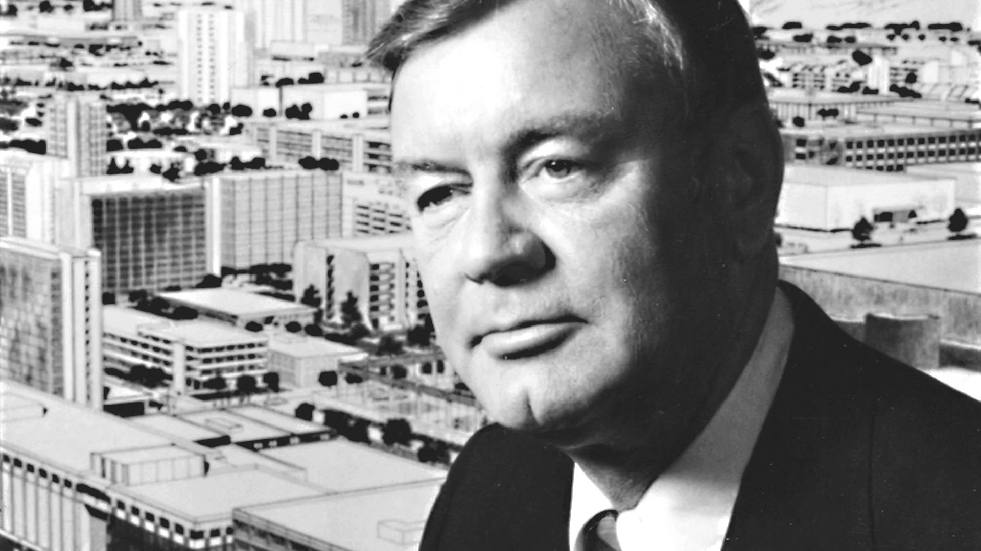 Black-and-white photo of a man with a serious expression, wearing a suit and tie, against a background of city buildings.