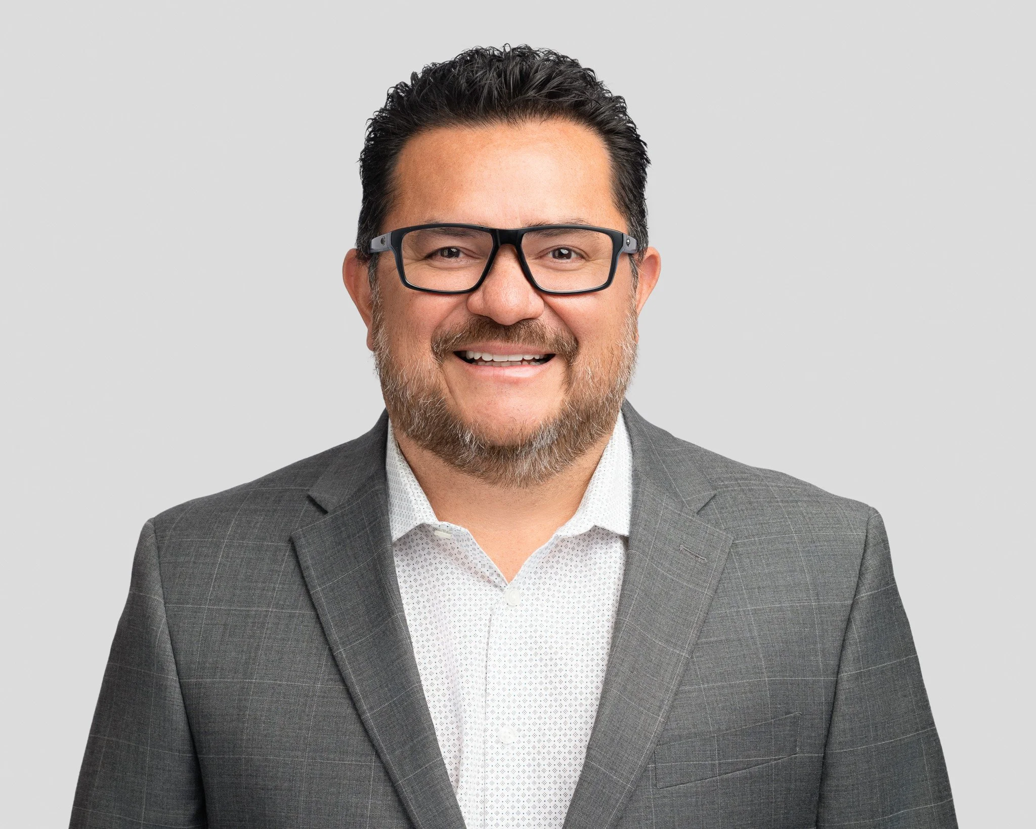 Headshot of a middle-aged man with dark hair, glasses, and a beard, wearing a gray suit and white dress shirt, smiling against a plain gray background.