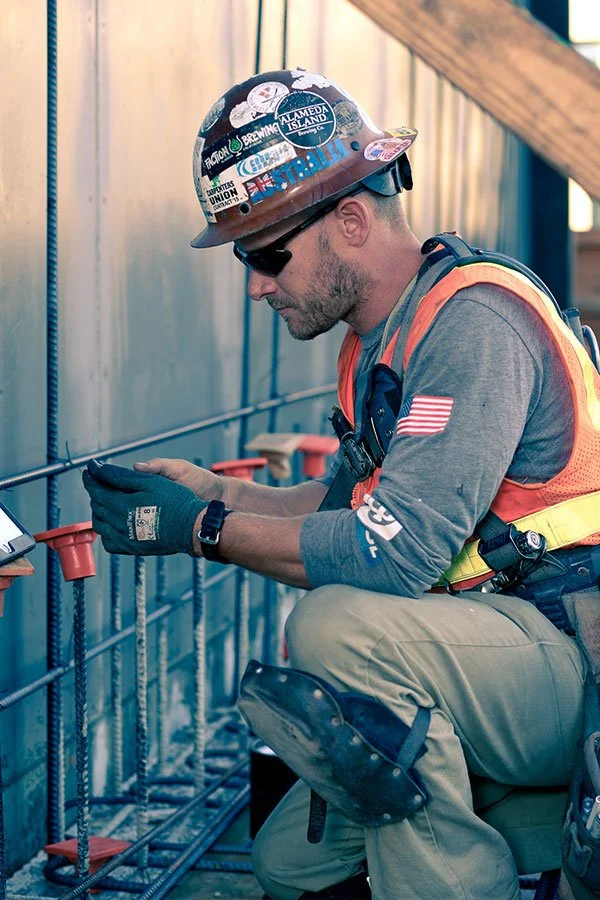 A construction worker wearing a safety helmet, sunglasses, and gloves is crouched down, examining a tablet or phone. He is dressed in a gray shirt with an American flag patch on the arm, an orange safety vest, and beige work pants, with construction tools and safety gear around him.