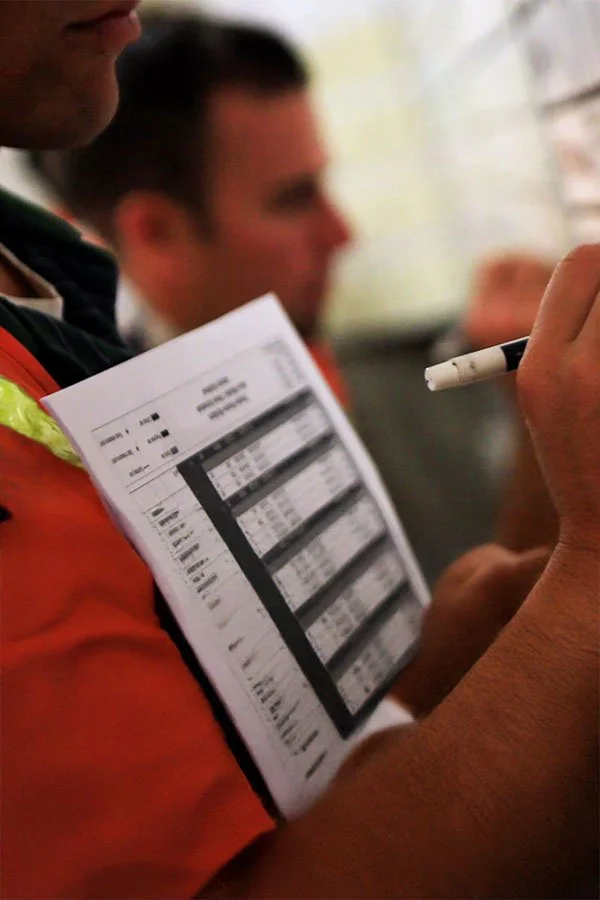 Close-up of a person wearing an orange safety vest and holding a clipboard with detailed charts or schedules, with another person blurred in the background.