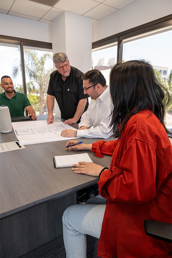 A group of five people in a meeting room, with blueprints and a notebook on the table. Two men are talking, one is standing and looking at the blueprints, the other is seated and writing in a notebook. A woman in a red jacket and white pants is seated in the foreground, taking notes. A man in a green shirt is seated smiling in the background. Large windows with trees and buildings outside are visible.
