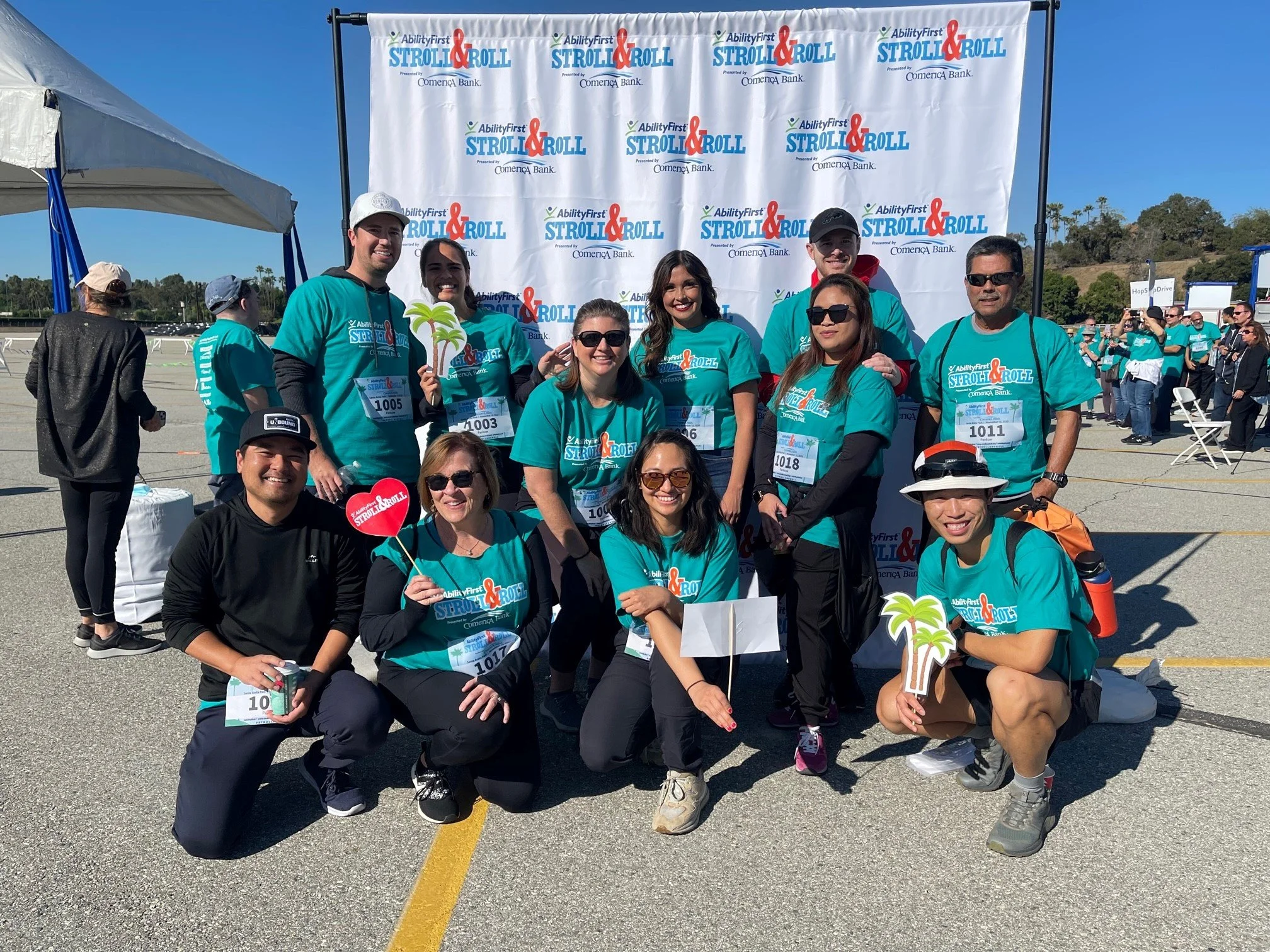 Group of people wearing teal shirts at a charity walk event called AbilityFirst Stroll & Roll, posing in front of a banner, some holding cutouts of palm trees and a heart with the event's logo.