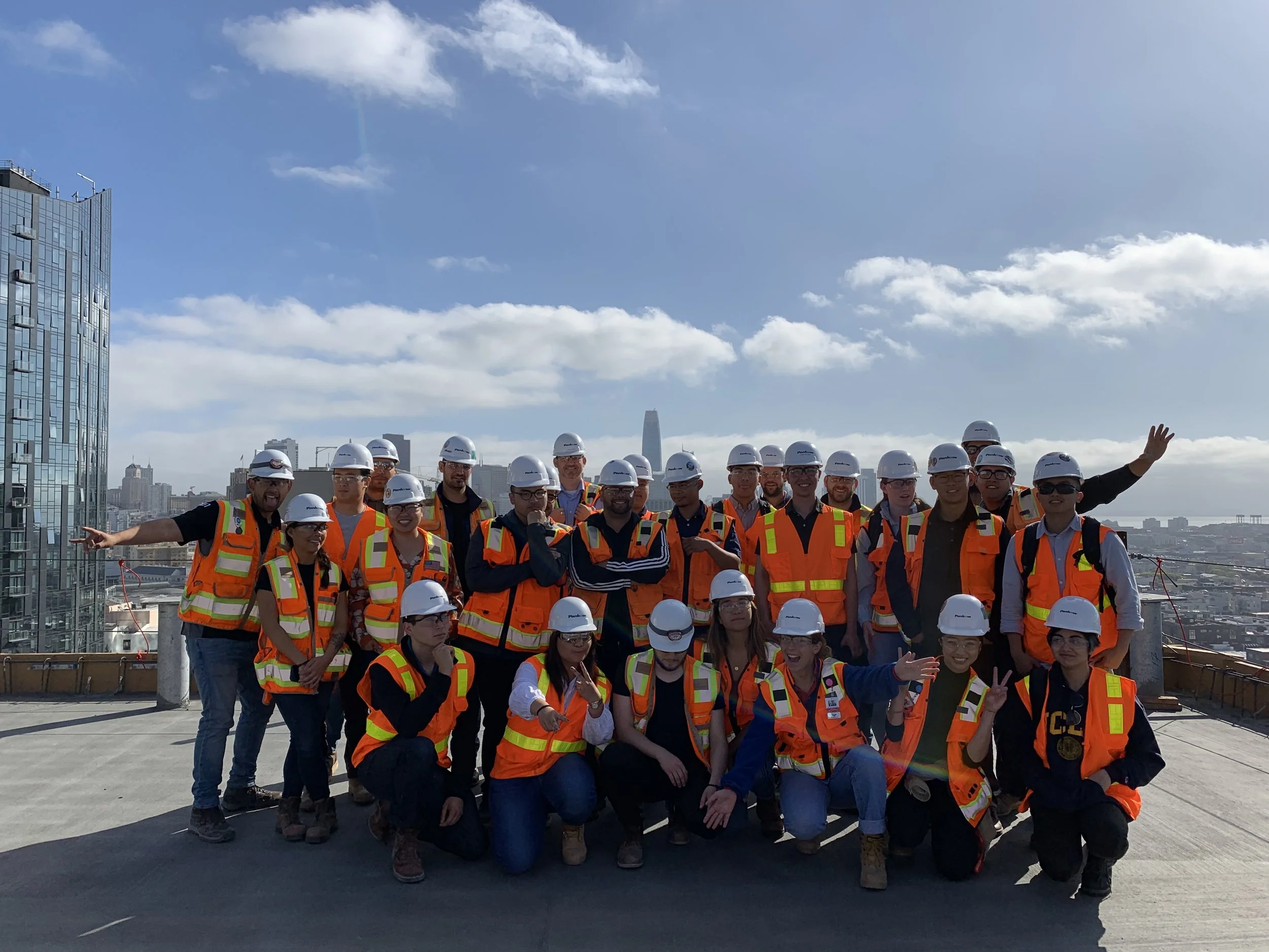 A large group of construction workers wearing orange safety vests and white safety helmets standing together on a high rooftop with a city skyline in the background, under a partly cloudy sky.
