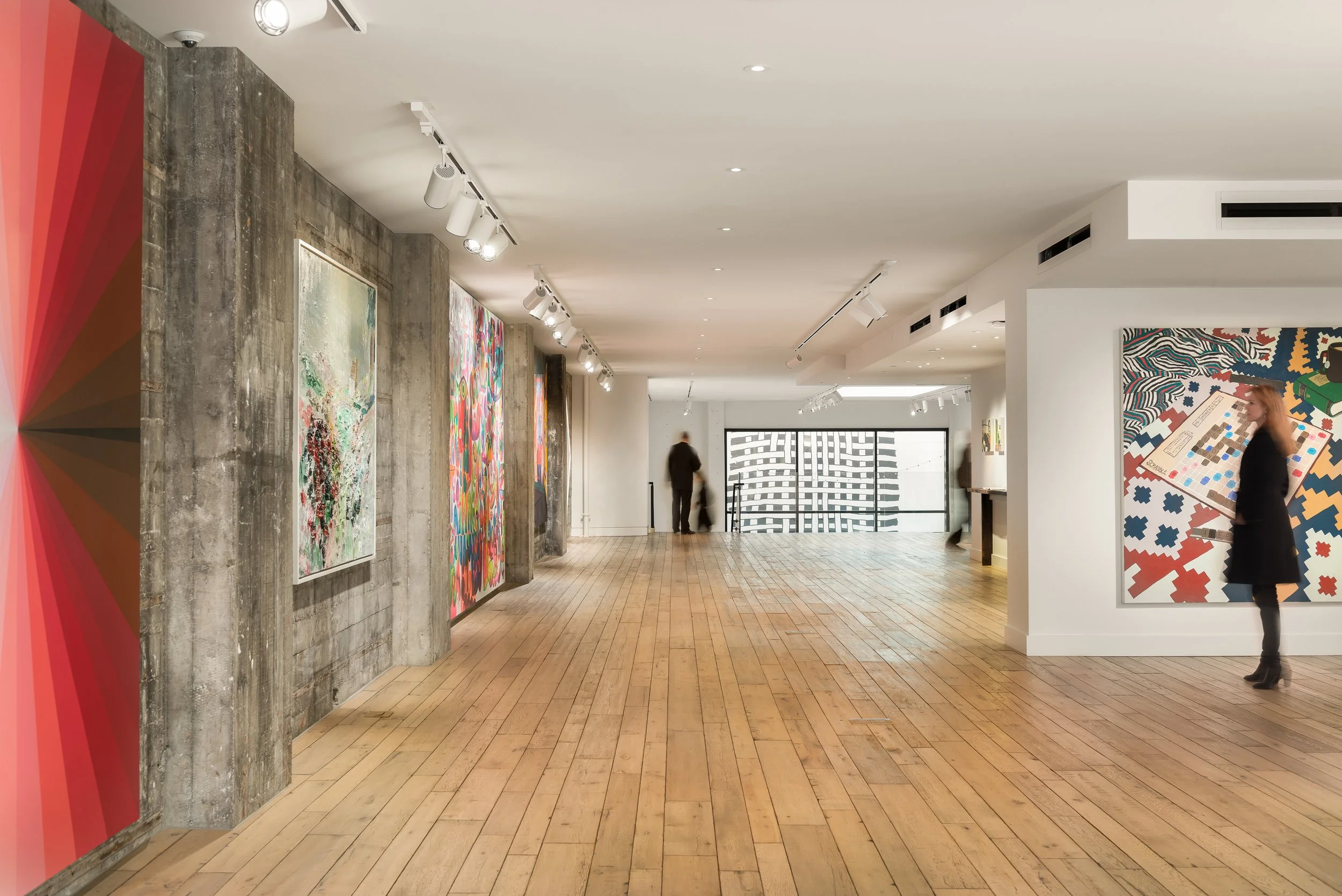 View of an art gallery with wooden floors, white walls, modern artwork, and track lighting, with a few visitors exploring the space.