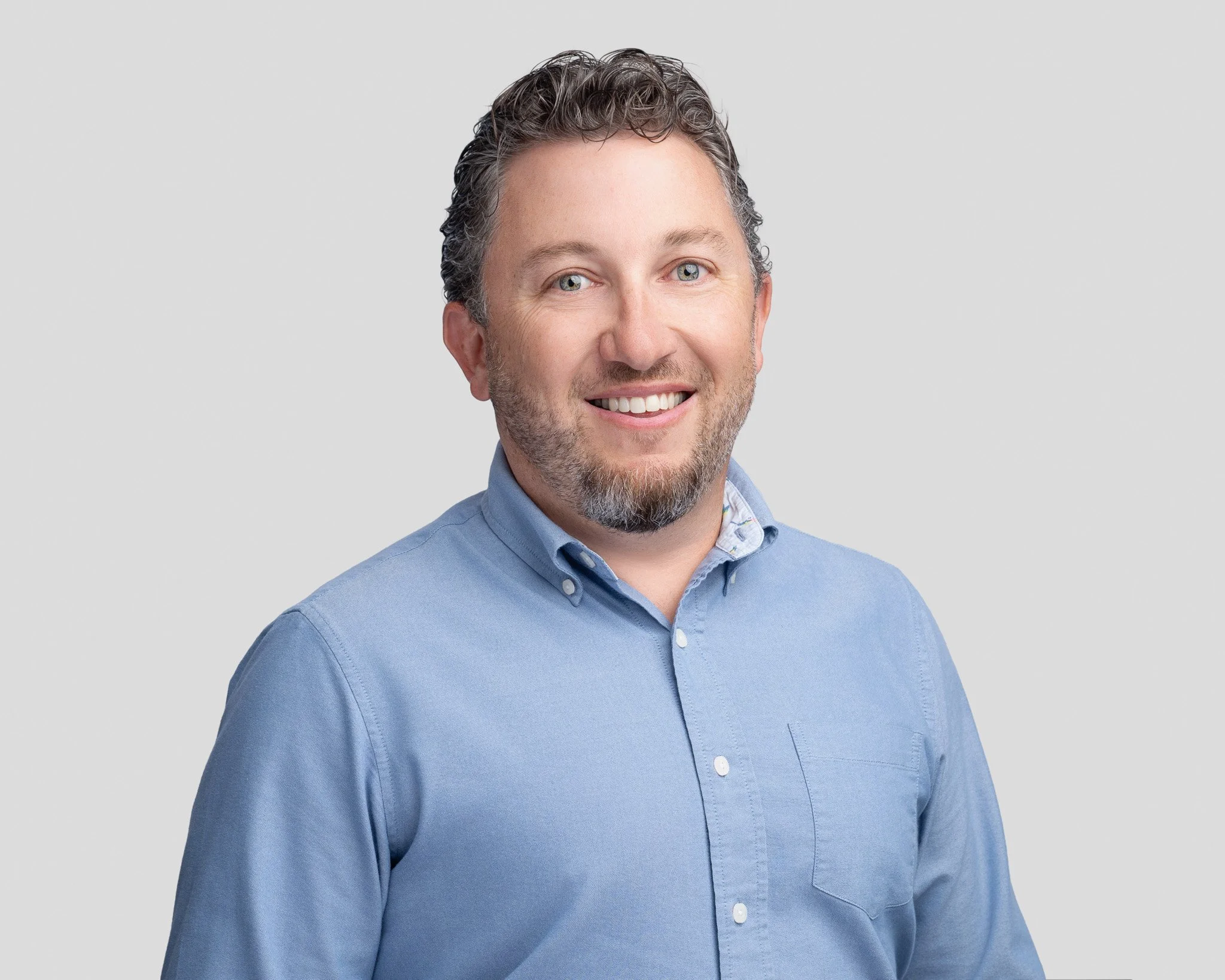 A smiling man with short, curly gray hair and a beard, wearing a light blue button-up shirt, standing against a plain light gray background.