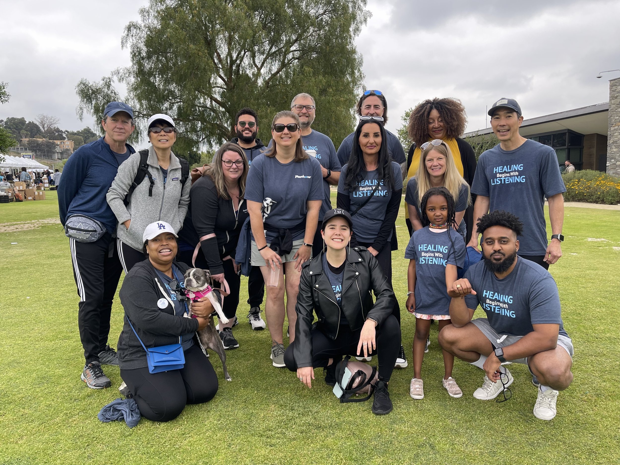 Group of 15 people, including children and adults, standing outdoors on grass field with trees and a building in the background. Some are wearing matching shirts with slogans like 'HEALING Begins With LISTENING'. One person is holding a small dog. It appears to be a community event or gathering.