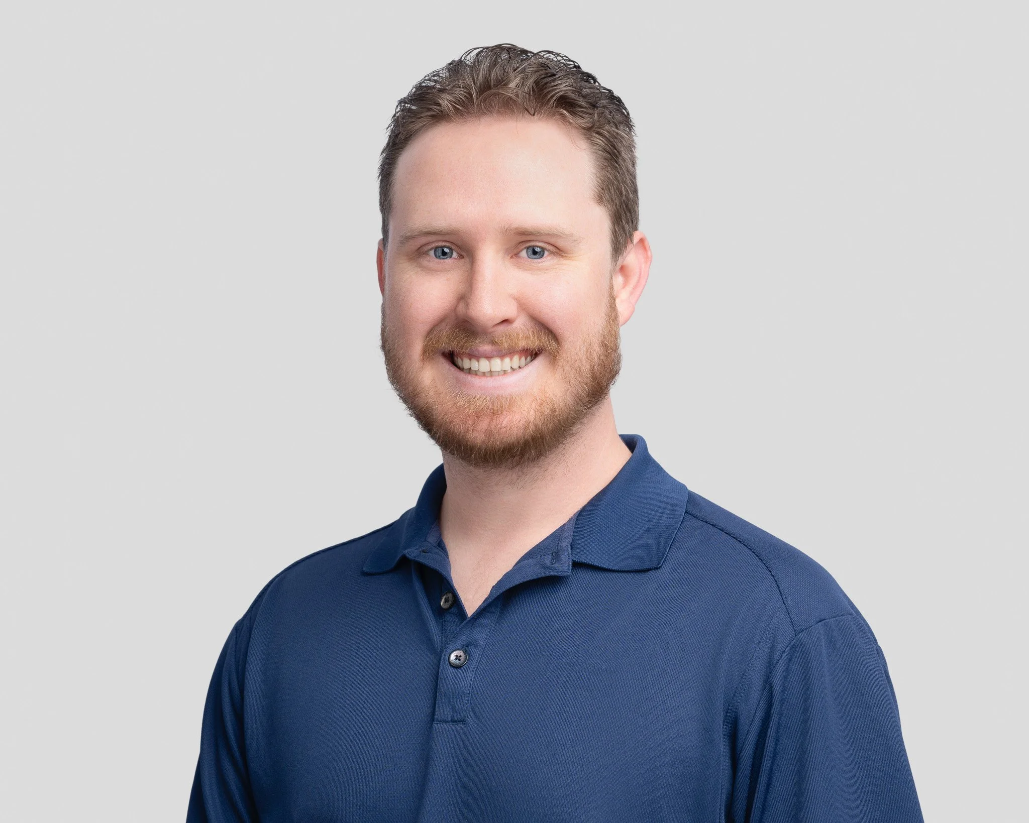A smiling man with a beard and blue eyes, wearing a navy polo shirt, against a plain light gray background.