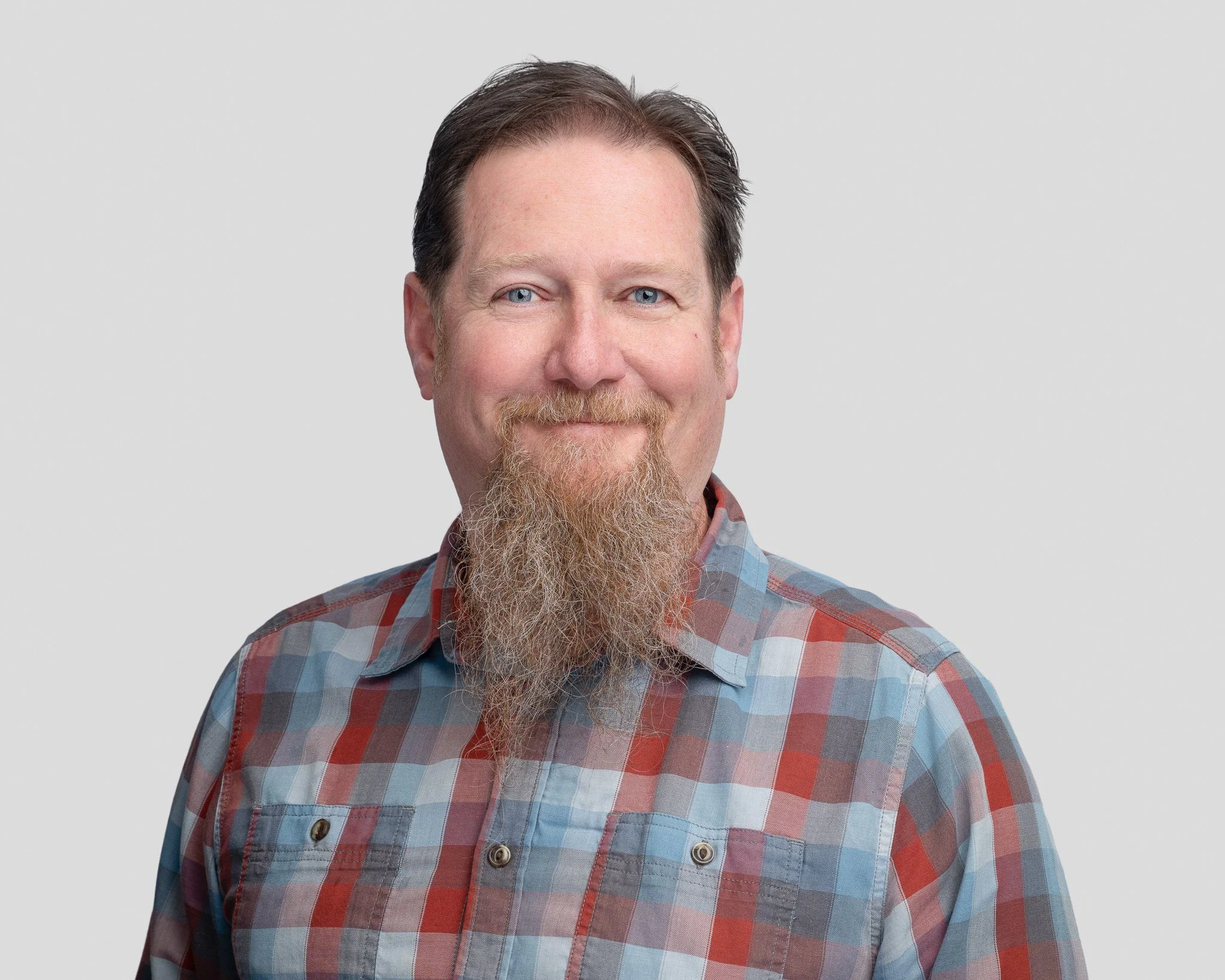 A man with a beard and light skin, smiling, wearing a red and blue plaid shirt against a plain background.