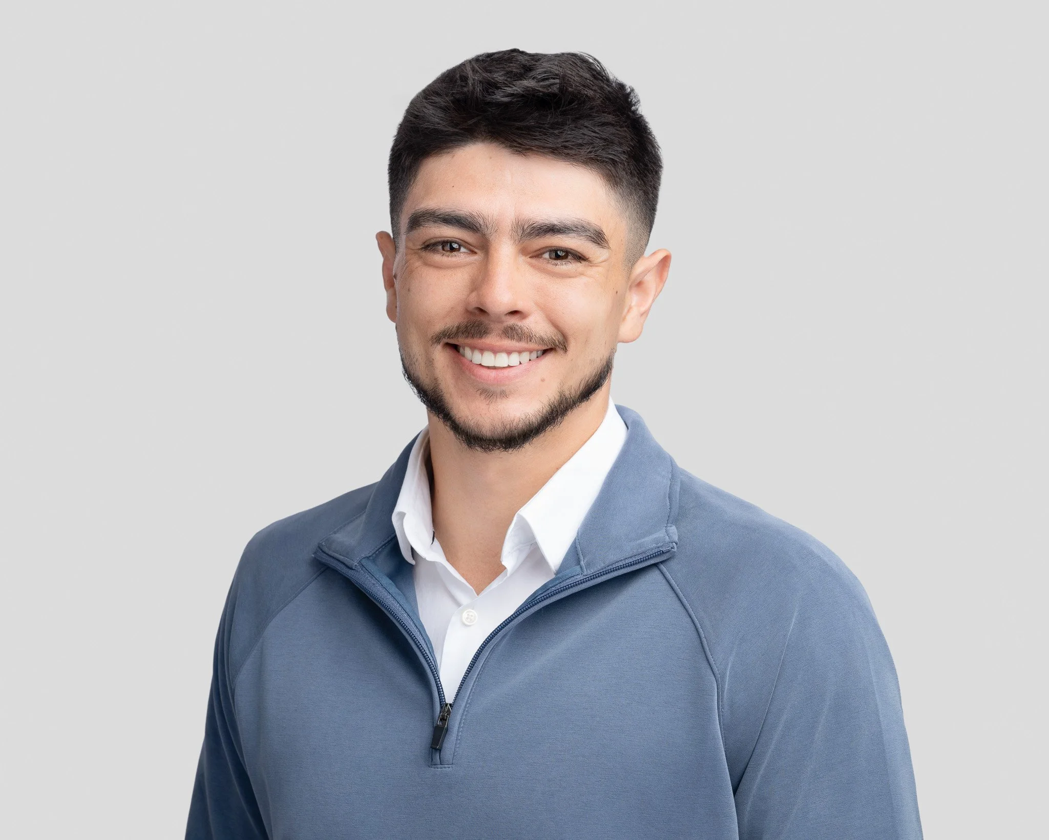 A young man with short dark hair, a beard, and a mustache, smiling, wearing a white collared shirt and a blue zip-up jacket, standing against a light gray background.