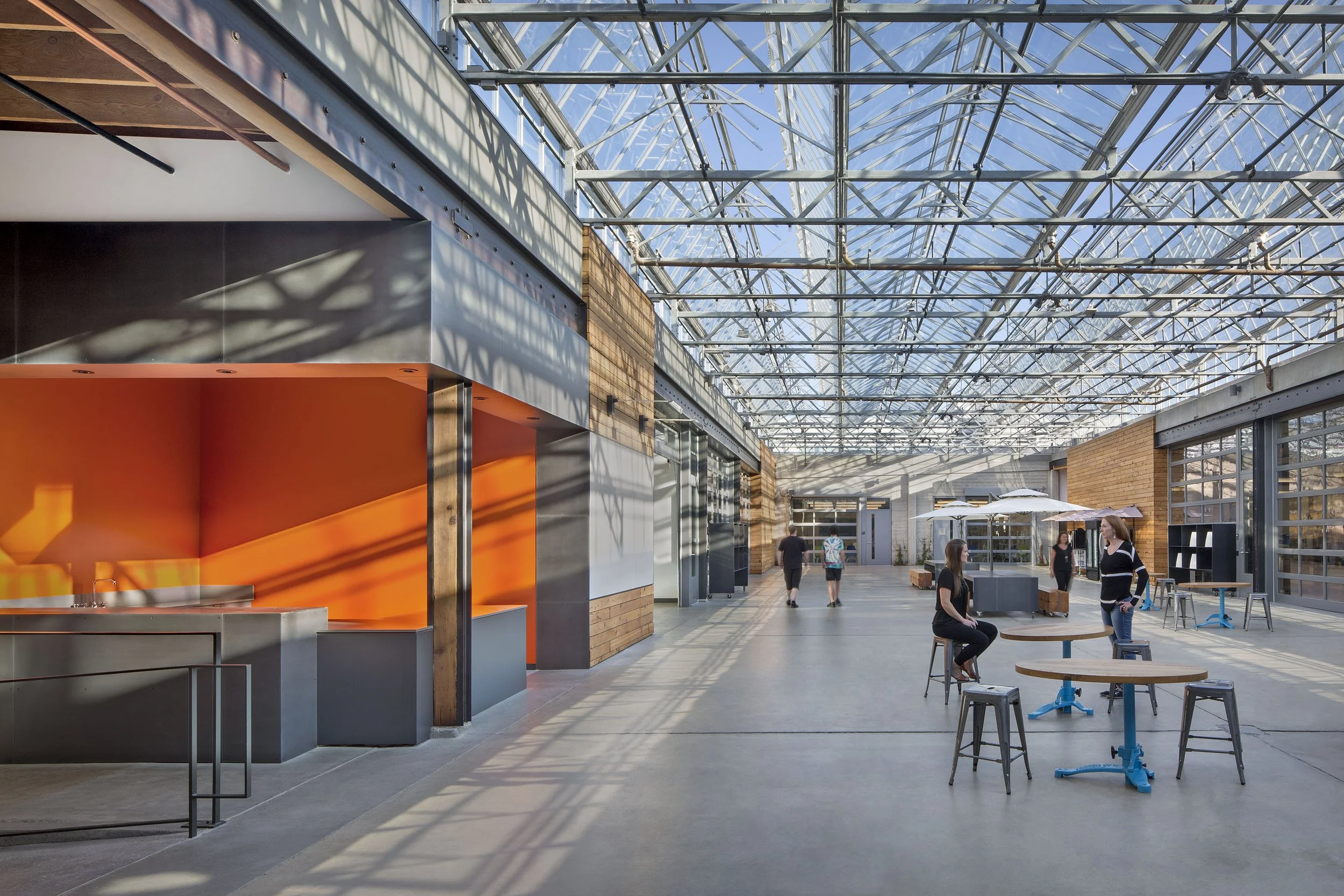 An interior or semi-outdoor space with a glass ceiling, showcasing modern design with brick and metal accents. Several people are present, including two women standing and talking at a high table, a woman sitting on a stool, and others walking in the