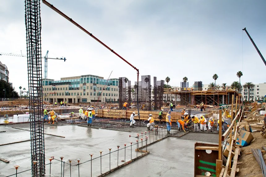 Construction site with workers pouring concrete for building foundation, cranes, and partially built structures, with modern buildings and palm trees in the background.