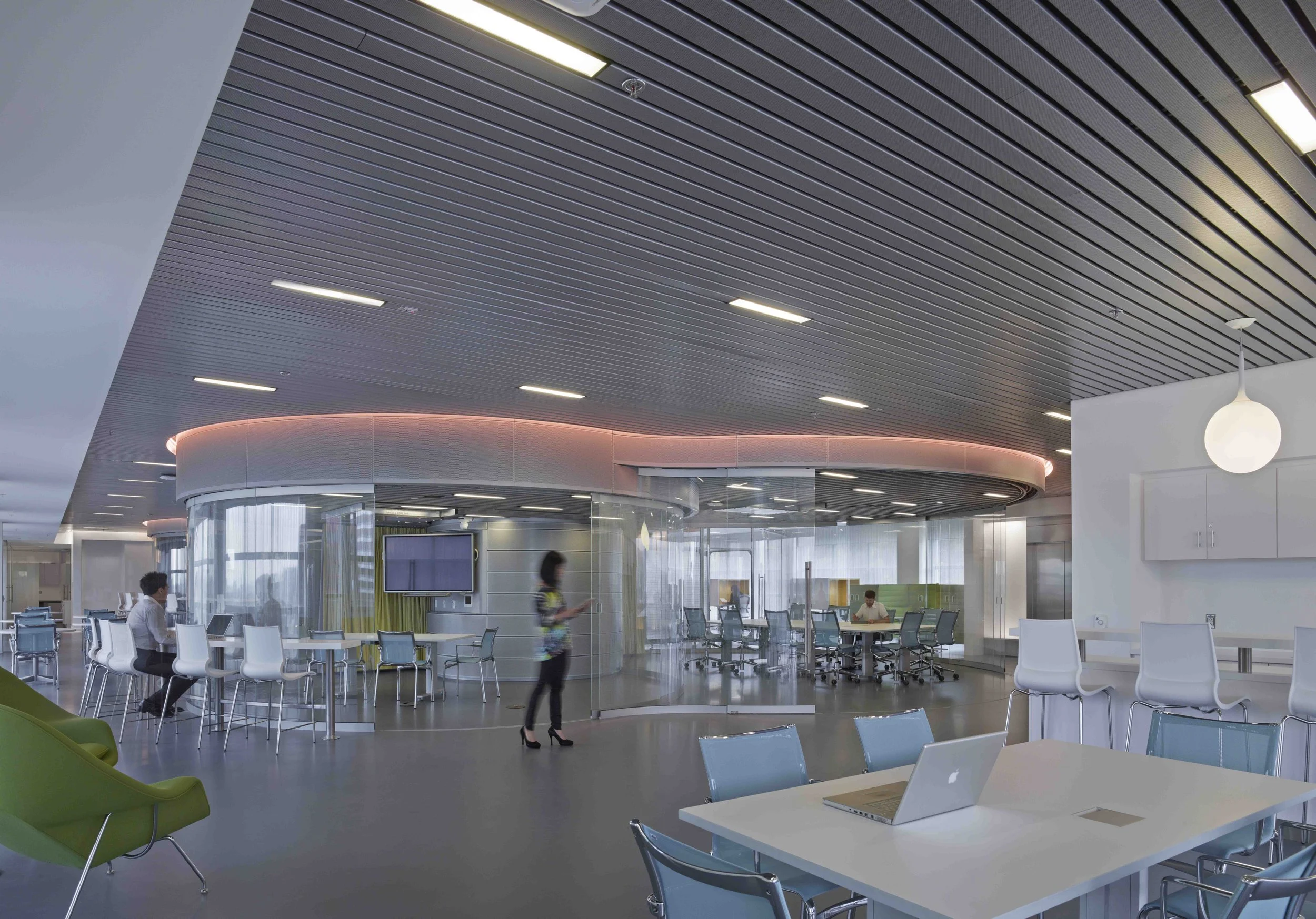 Modern office space with glass partitions, white chairs, a green lounge chair, and a table with an open laptop. People are working and walking, with a ceiling featuring black slats and built-in lighting.