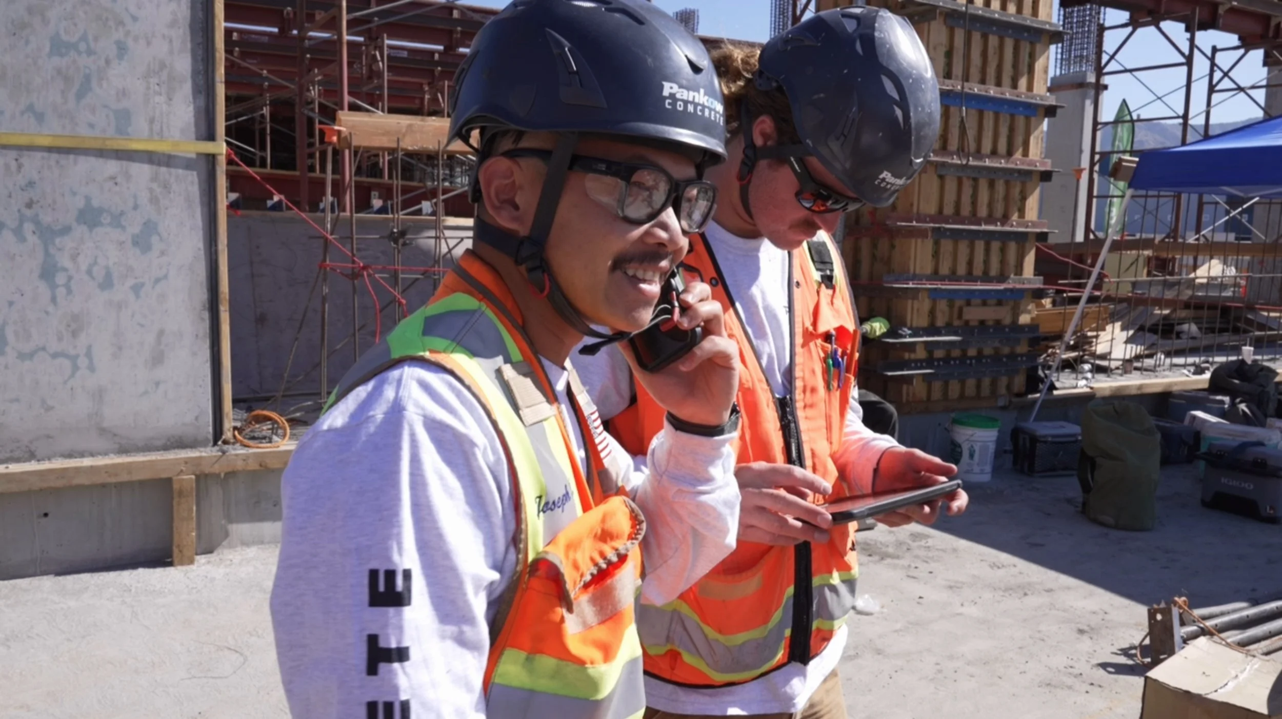 Two construction workers wearing safety helmets and reflective vests are on a construction site. One is talking on a walkie-talkie, while the other is looking at a tablet.