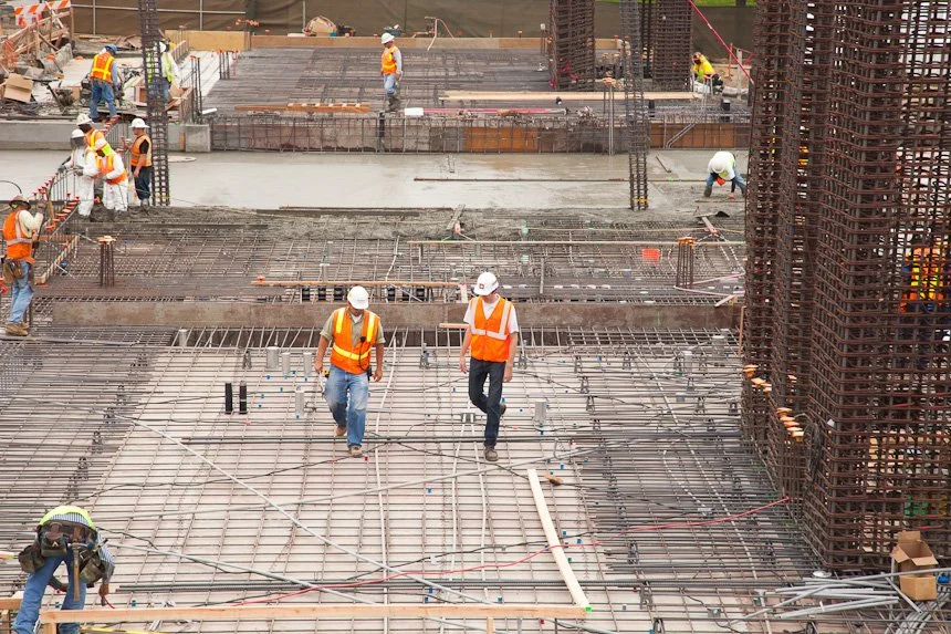Construction workers wearing orange safety vests and white hard hats working on a building site with steel rebar and concrete foundation.