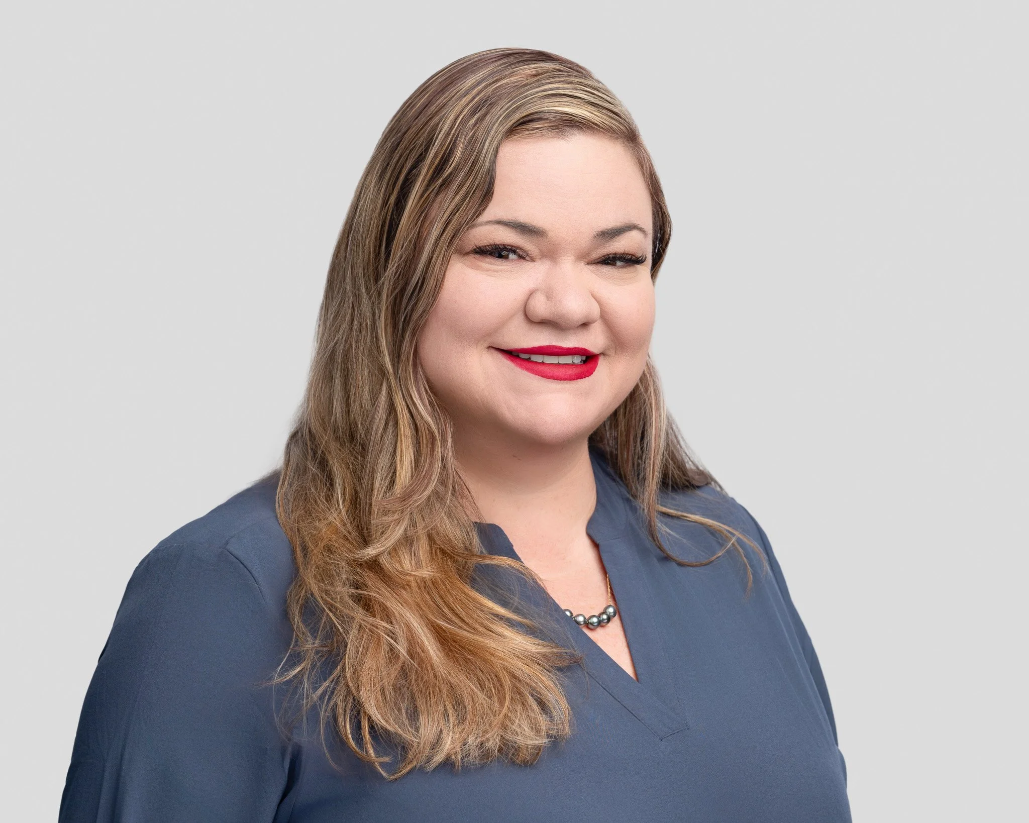 Portrait of a woman with long wavy hair wearing a navy blue top and a silver necklace, smiling against a plain gray background.