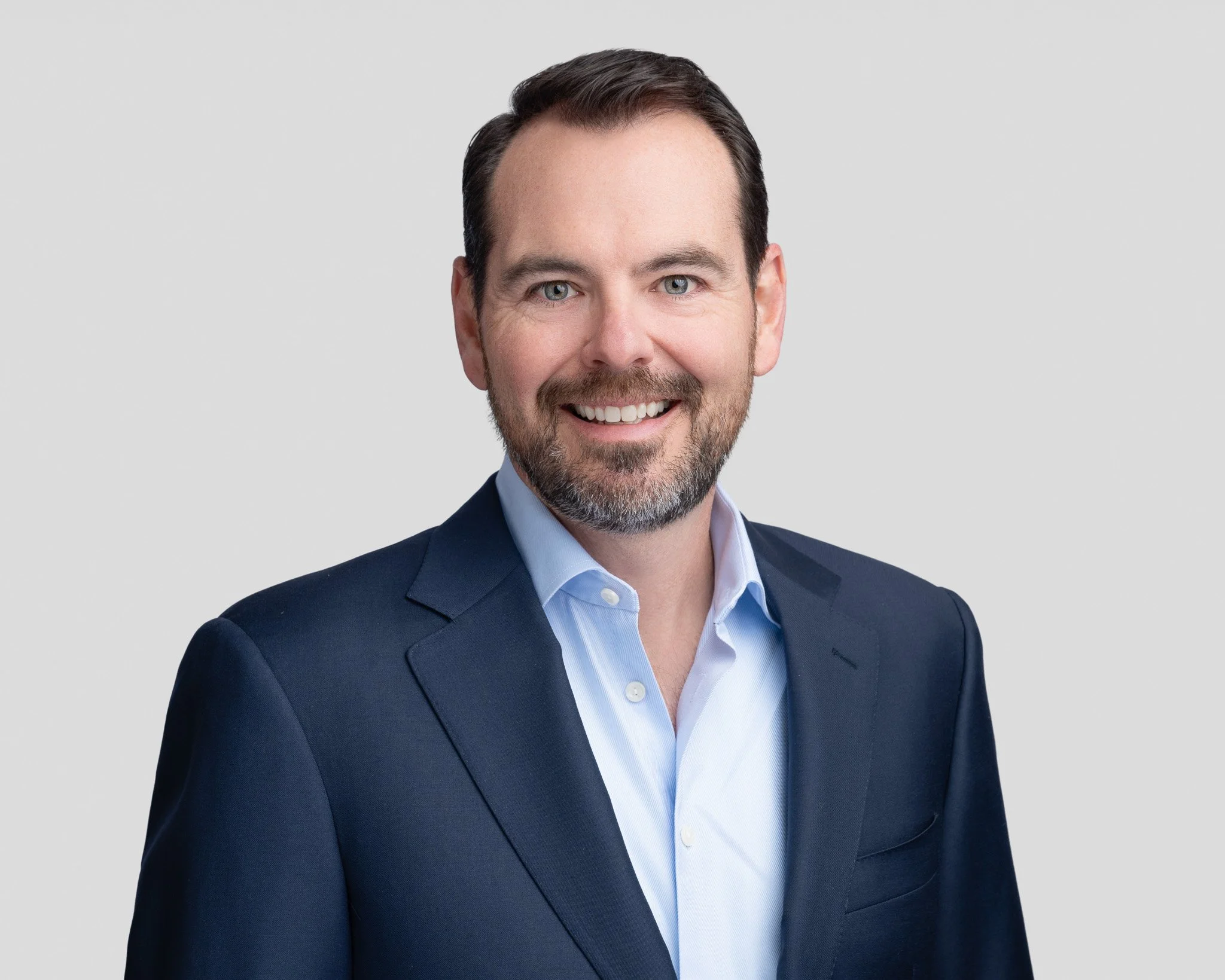 Professional headshot of a smiling man with dark hair and beard, wearing a navy suit and light blue shirt, against a plain gray background.