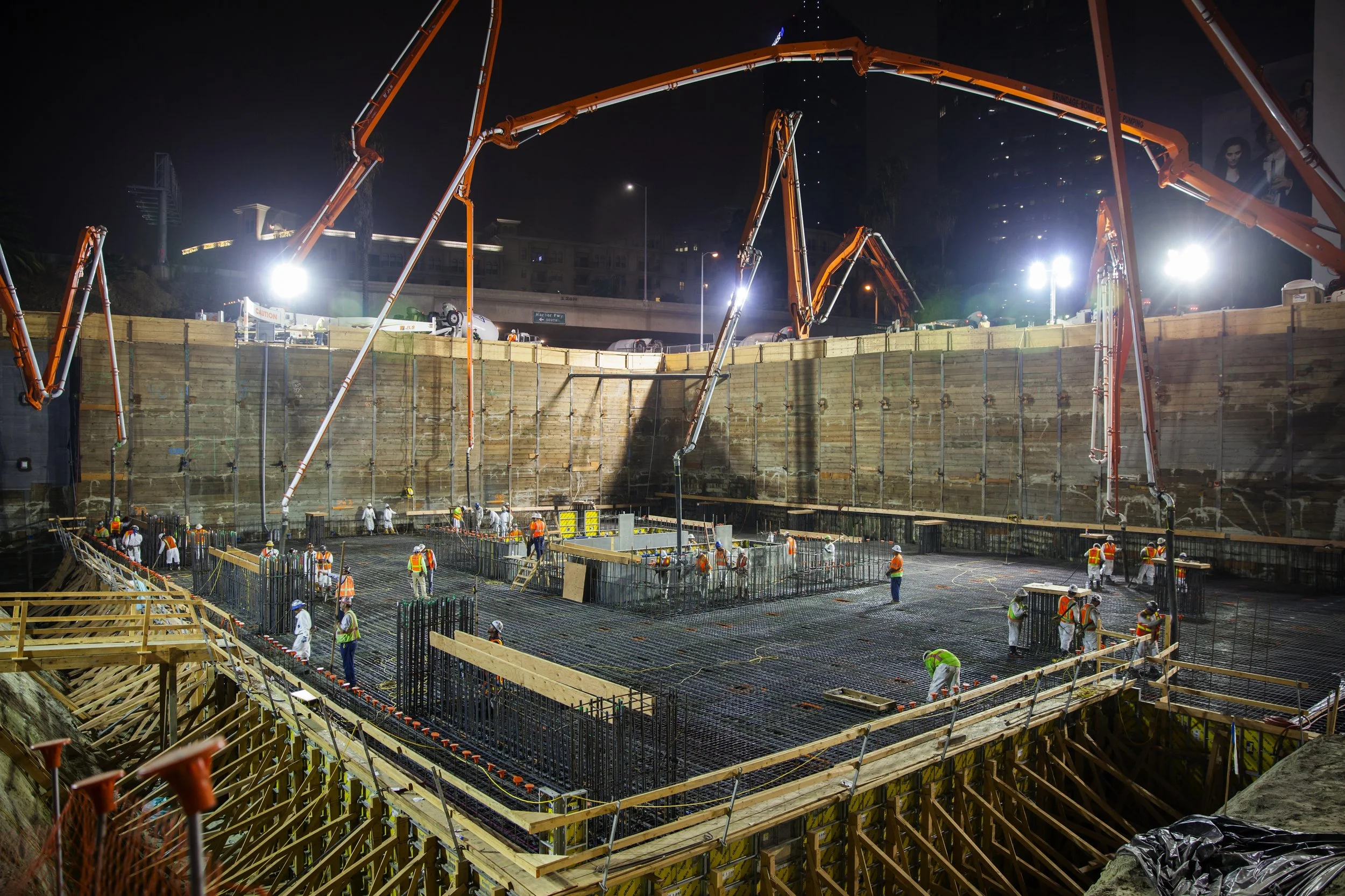 Construction workers at night working on a building foundation with large construction cranes and wooden formwork.